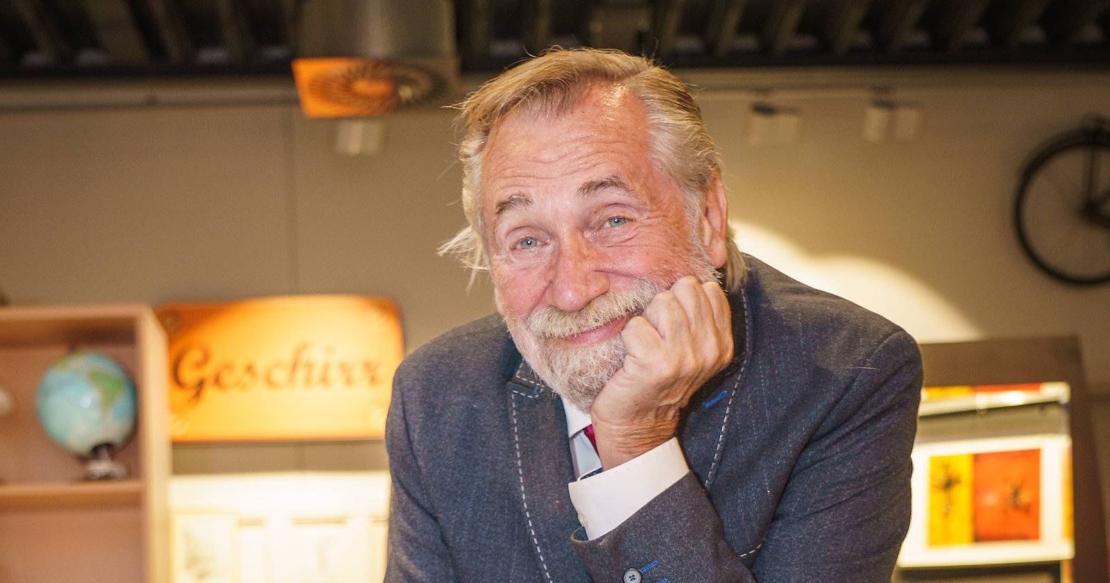 Porträt von Peter Rapp mit grauem Bart und Anzug, lächelnd, den Kopf in die Hand gestützt, im Hintergrund ein Schild mit der Aufschrift „Geschirr“.