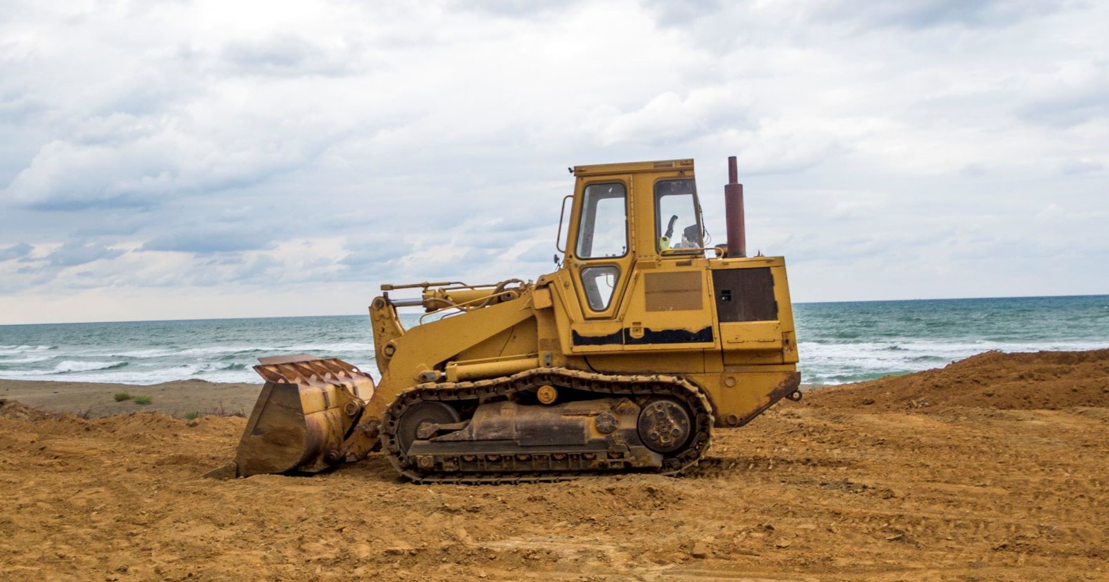 Gelbe Planierraupe auf sandigem Strandabschnitt vor dem Meer, unter bewölktem Himmel.