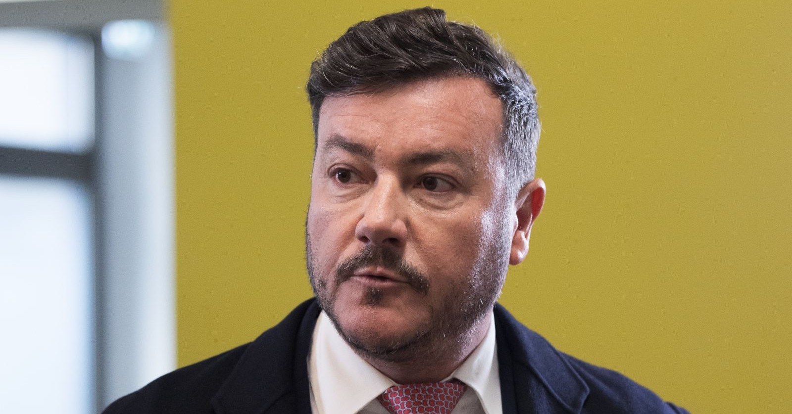 René Benko blickt ernst zur Seite, im Hintergrund eine gelbe Wand.