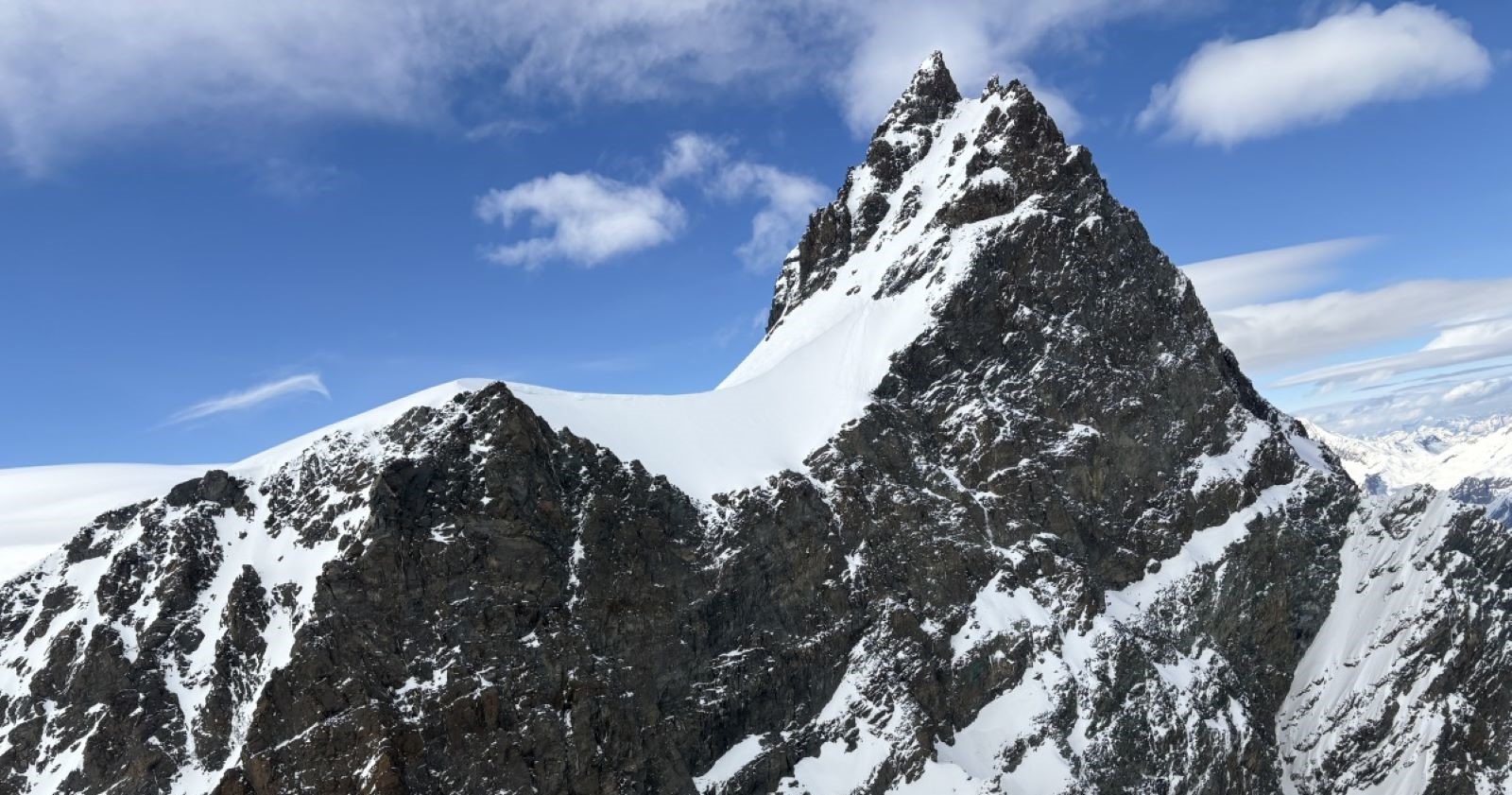Verschneiter, steil aufragender Gipfel des Rimpfischhorns in den Walliser Alpen unter blauem Himmel mit vereinzelten Wolken. Links davon ein schneebedeckter Sattel, rechts steile Felsflanken.