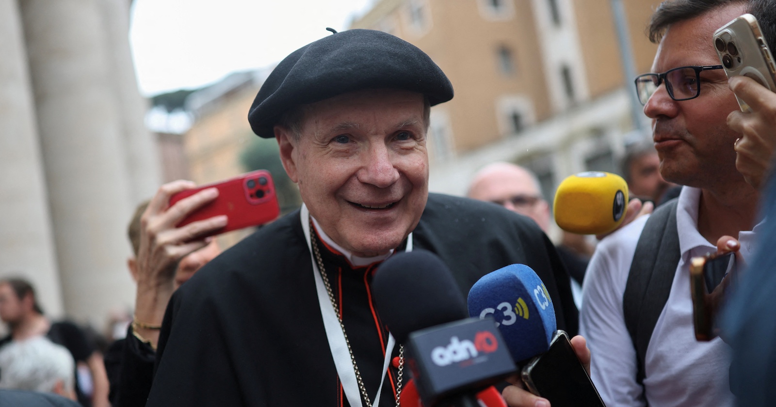 Kardinal Christoph Schönborn, freundlich lächelnd mit schwarzem Barett und rotem Kardinalsrang, wird von mehreren Journalisten mit Mikrofonen und Smartphones umringt. Im Hintergrund sind Menschen und Gebäude in Rom zu sehen.