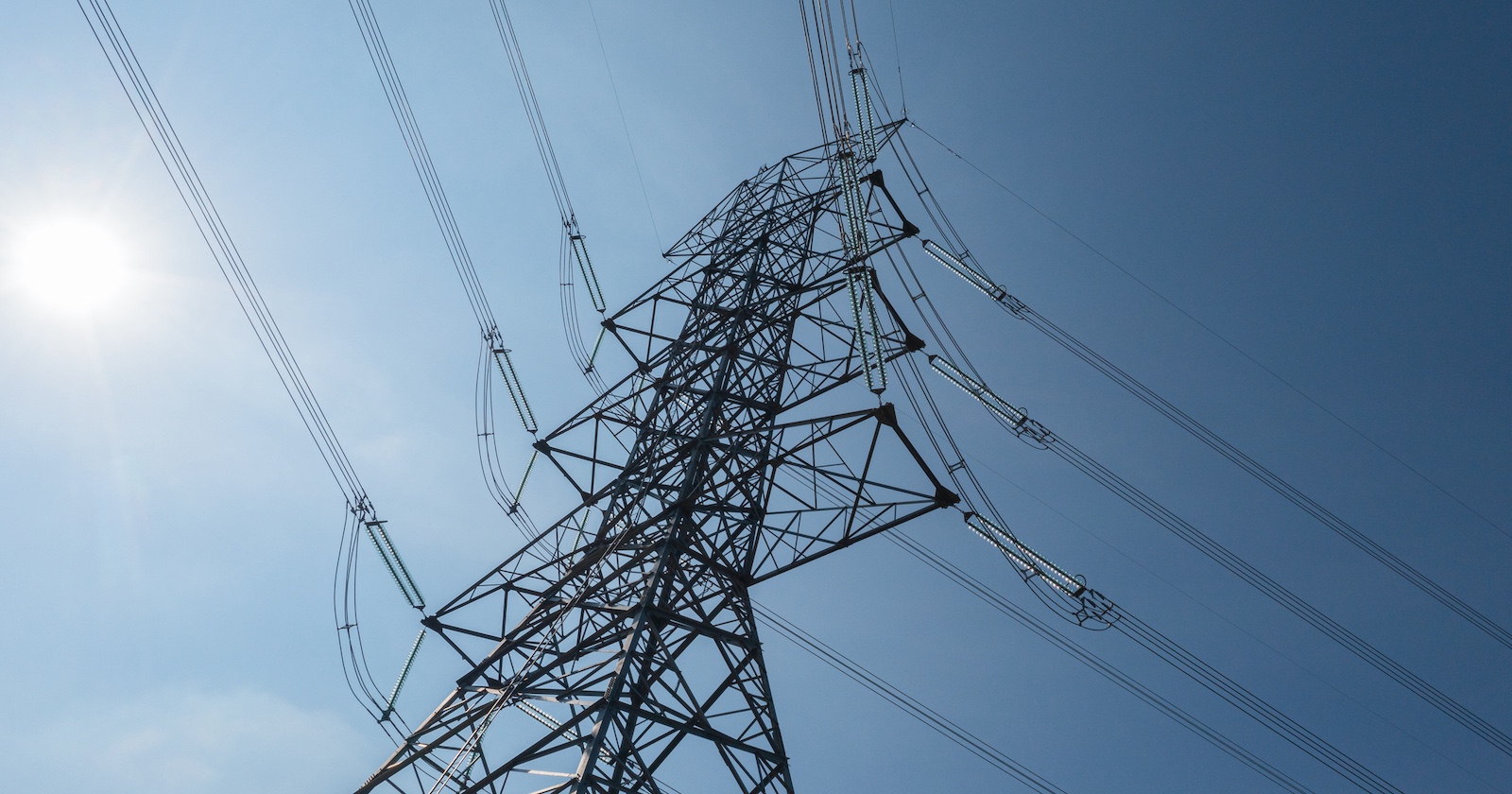 Hochspannungsmast mit mehreren Stromleitungen vor blauem Himmel und strahlender Sonne – Symbolbild für Stromversorgung.