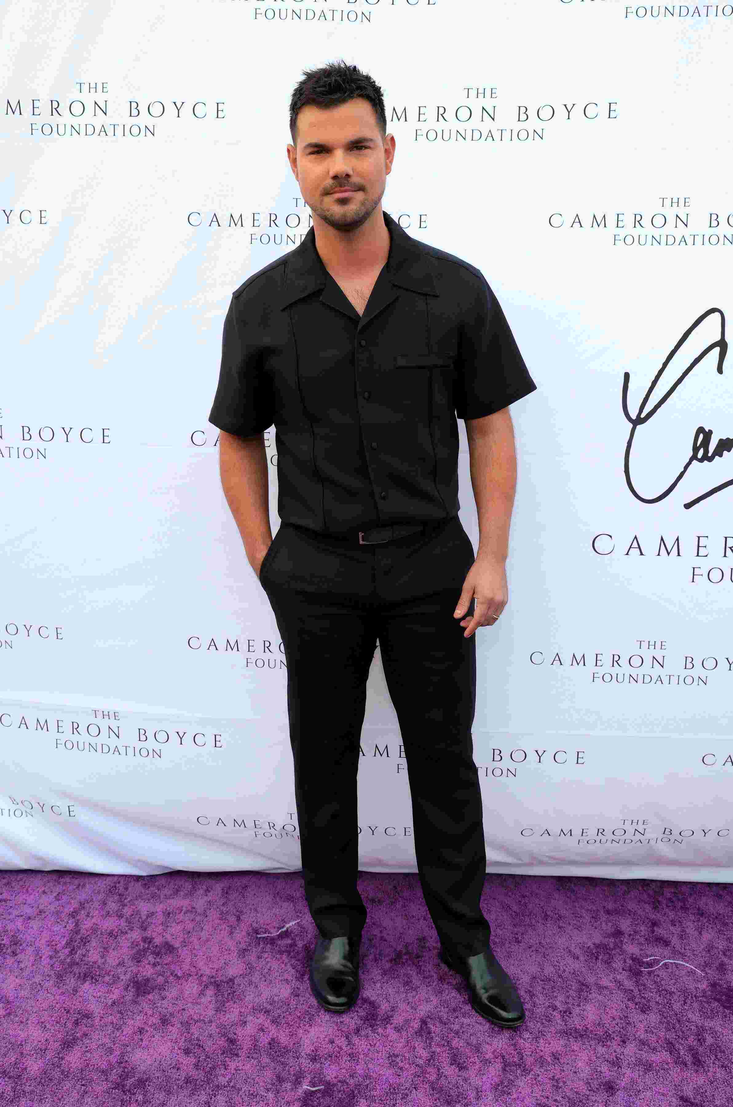 Taylor Lautner mit schwarzer Hose, schwarzem Hemd steht lässig auf einem violetten Teppich mit einer Hand in der Hosentasche.