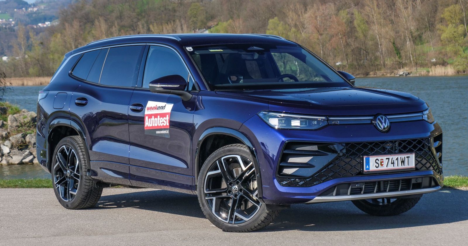 Ein lila VW Tayron parkt am Ufer eines Gewässers. Am anderen Ufer ist ein dichter Laubwald zu sehen.