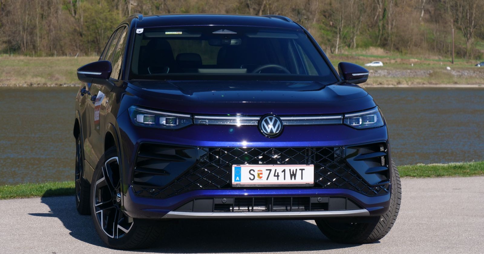 Ein lila VW Tayron - der in der Sonne leuchtet - steht am Ufer eines Gewässers. Am anderen Ufer sind Bäume zu sehen, die an einem Hang wachsen.