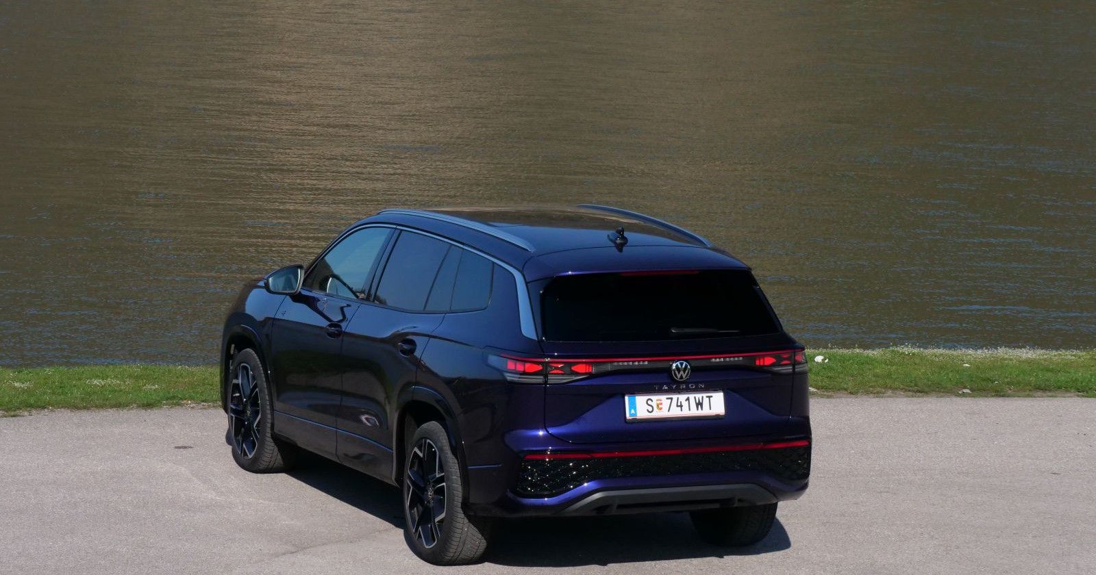 Ein lila VW Tayron, dessen Heck man sieht, parkt am Ufer eines Gewässers. Zwischen Wasser und Asphalt ist ein schmaler Streifen Gras zu sehen.