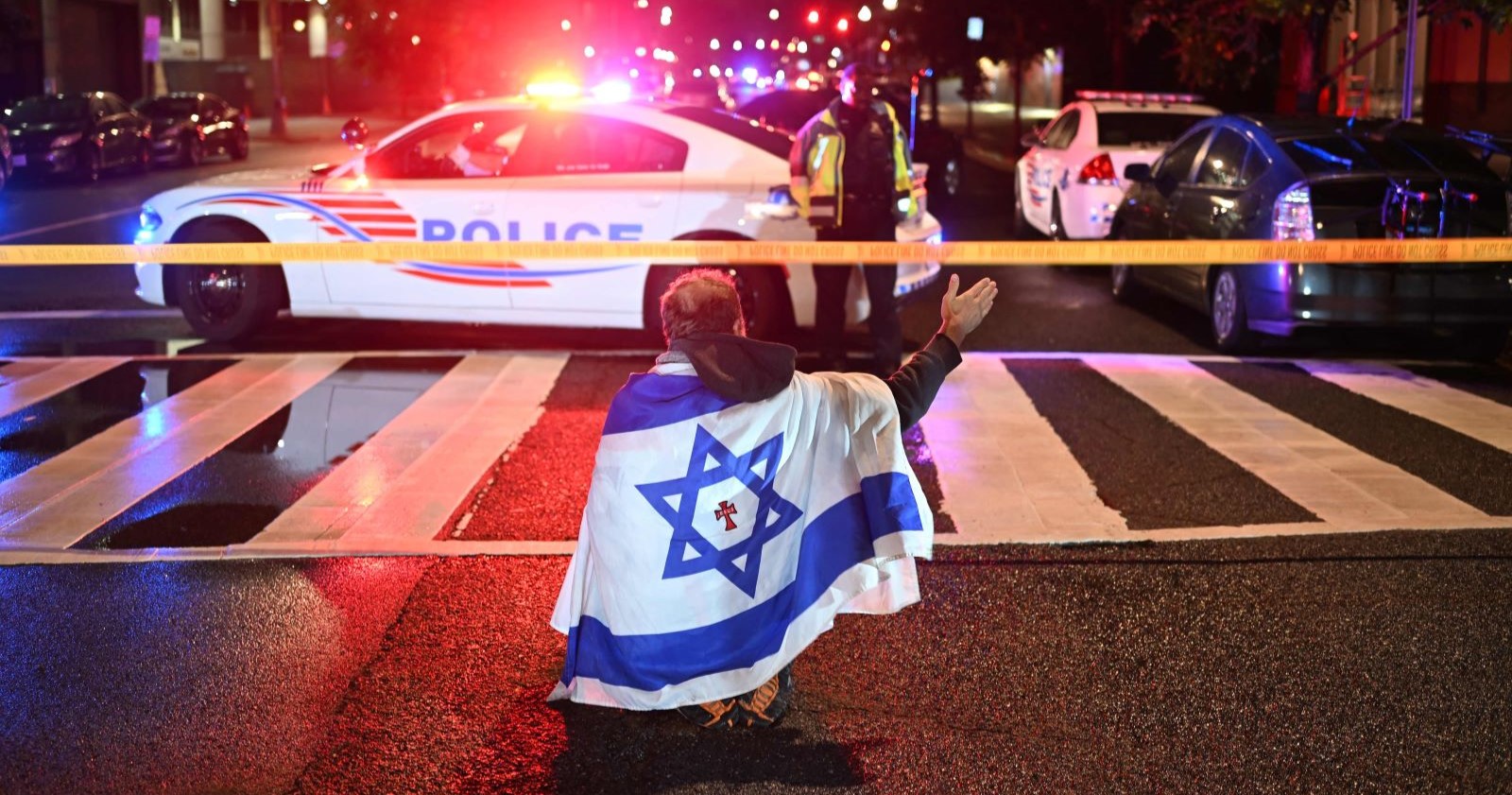 Ein Mann mit einer israelischen Flagge auf dem Rücken kniet nachts auf einer gesperrten Straße vor einem mit Blaulicht beleuchteten Polizeiwagen in Washington D.C. – Tatort eines tödlichen Anschlags auf israelische Botschaftsmitarbeiter.