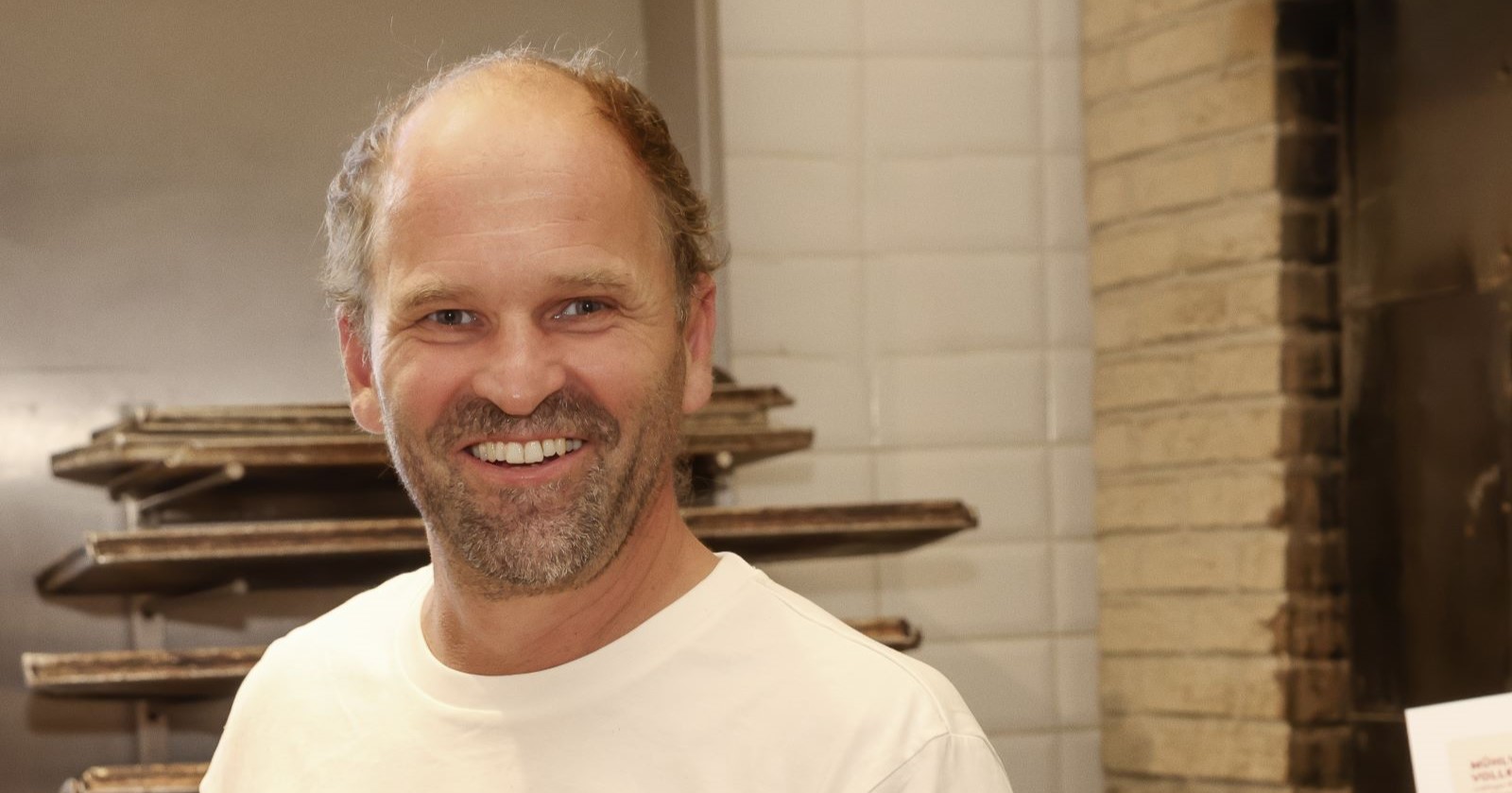 Helmut Gragger: Ein lachender Mann mit kurzem Haar und Bart, gekleidet in ein weißes T-Shirt mit der Aufschrift „Holzofenbäckerei Gragger“, hält in einer Bäckerei drei runde Laibe frisches Brot auf einem Holzbrett. Im Vordergrund sind Croissants und Käsegebäck zu sehen, im Hintergrund ein gemauerter Ofen.