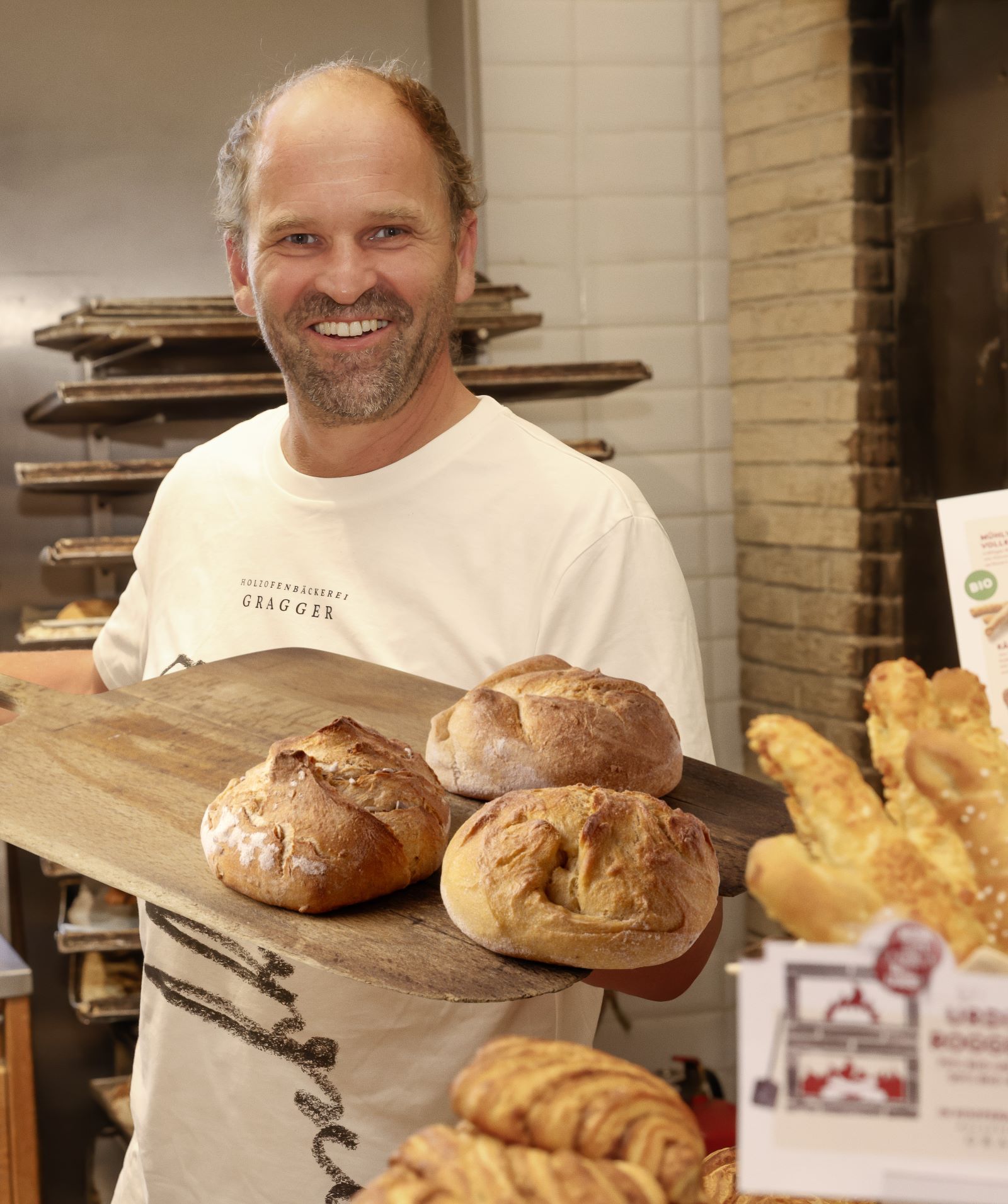 Ein lachender Mann mit kurzem Haar und Bart, gekleidet in ein weißes T-Shirt mit der Aufschrift „Holzofenbäckerei Gragger“, hält in einer Bäckerei drei runde Laibe frisches Brot auf einem Holzbrett. Im Vordergrund sind Croissants und Käsegebäck zu sehen, im Hintergrund ein gemauerter Ofen.
