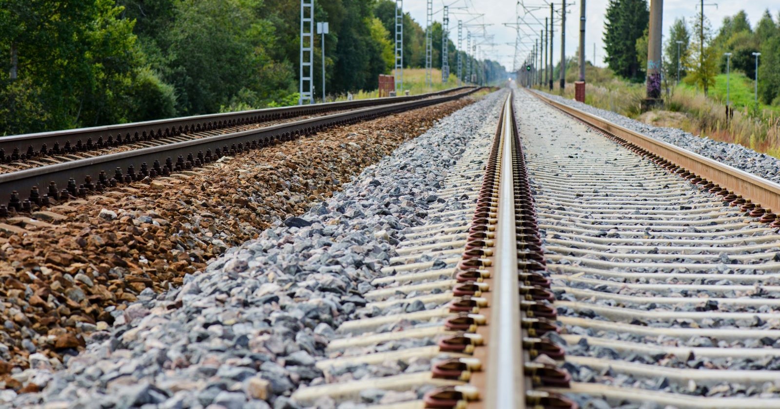Zwei parallel verlaufende Bahngleise mit Schotterbett und Oberleitungen, aufgenommen bei Tageslicht in einer ländlichen Gegend.