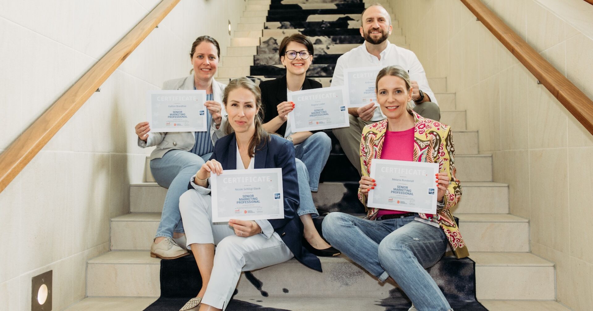 Absolventen des &quot;Senior Marketing Professional&quot;-Programms sitzen mit ihren Urkunden auf einer Stiege