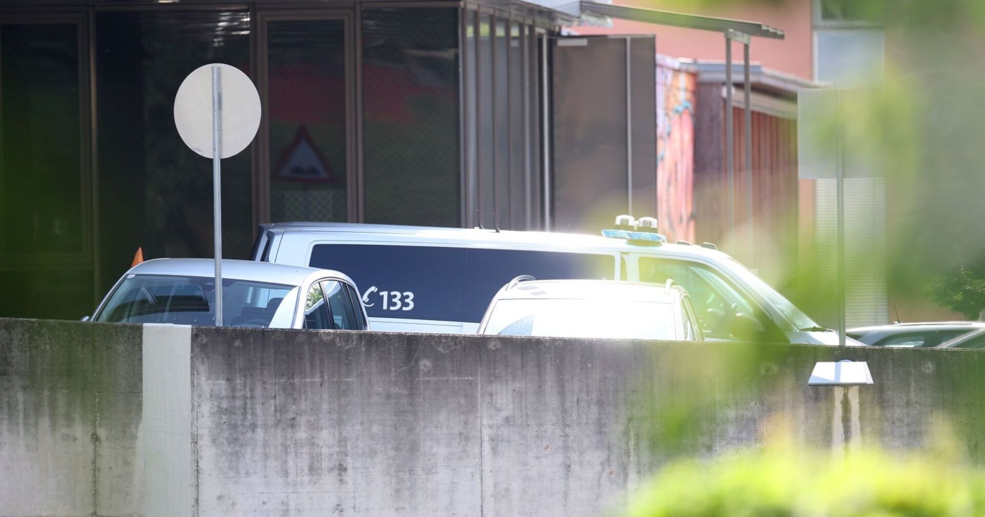 Polizeifahrzeuge stehen vor einem Wohngebäude hinter einer Mauer, Nummer 133 sichtbar, Szene abgeschirmt und teilweise von Grün verdeckt.