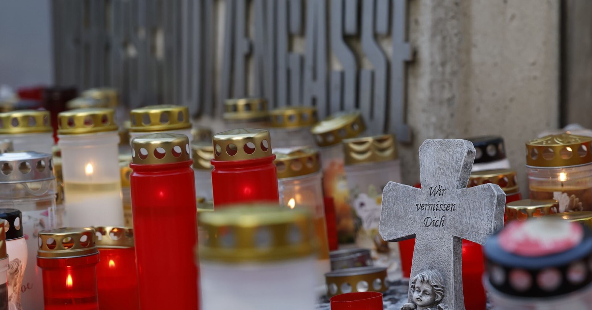 Zahlreiche rote und weiße Grablichter stehen vor einer Wand mit dem Schriftzug „Dreierschützengasse“. Im Vordergrund ein graues Steinkreuz mit der Inschrift „Wir vermissen Dich“ und einer Engelsfigur.