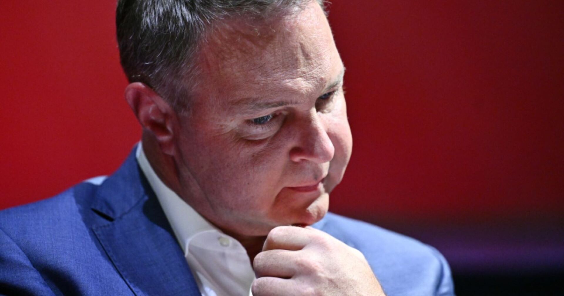 Andreas Babler in einem blauen Anzug vor rotem Hintergrund, mit nachdenklichem Gesichtsausdruck und der Hand am Kinn.