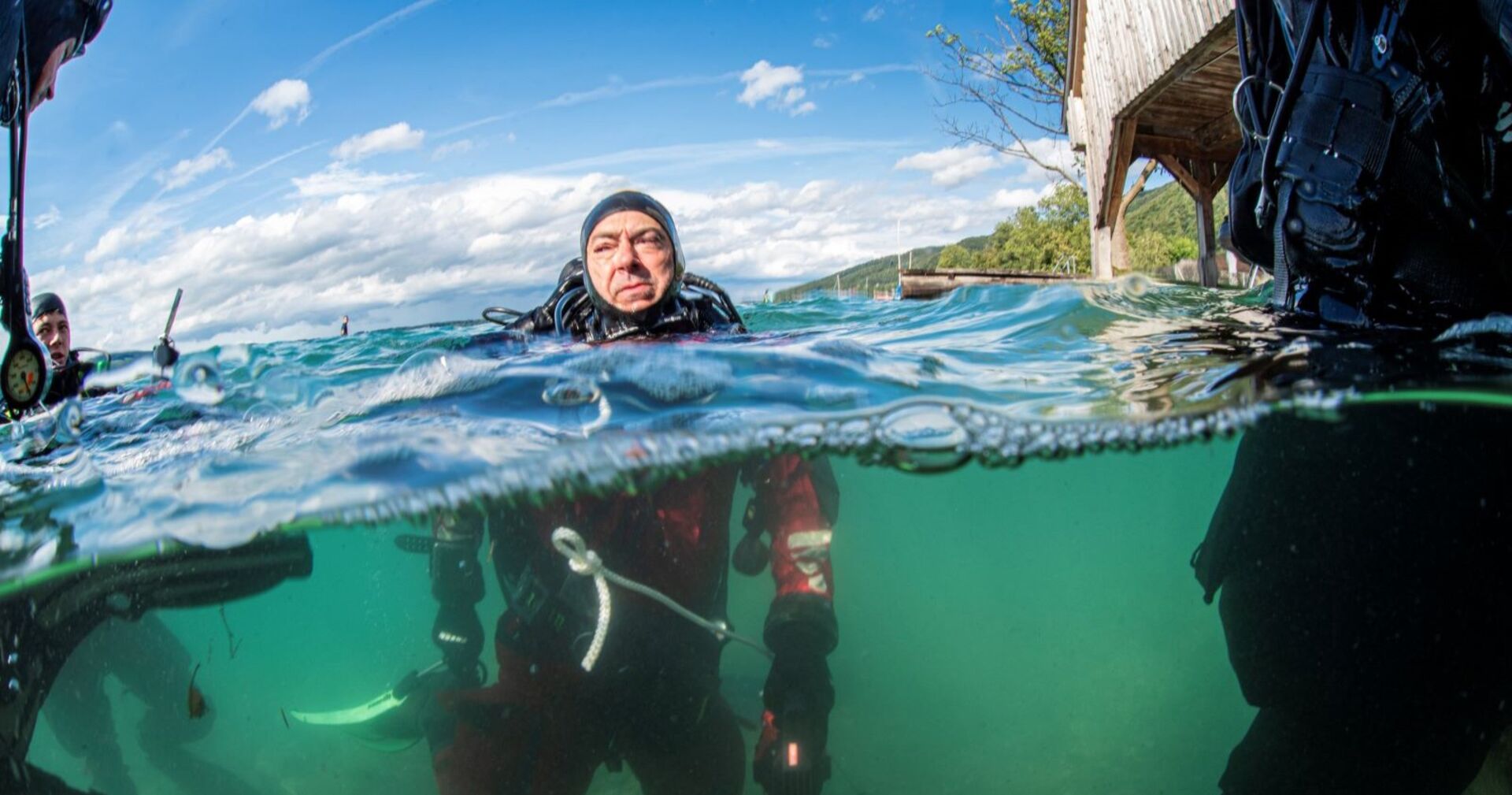Taucher in voller Ausrüstung im Attersee, teilweise unter Wasser fotografiert, mit Blick auf einen Holzsteg und das Ufer im Hintergrund.