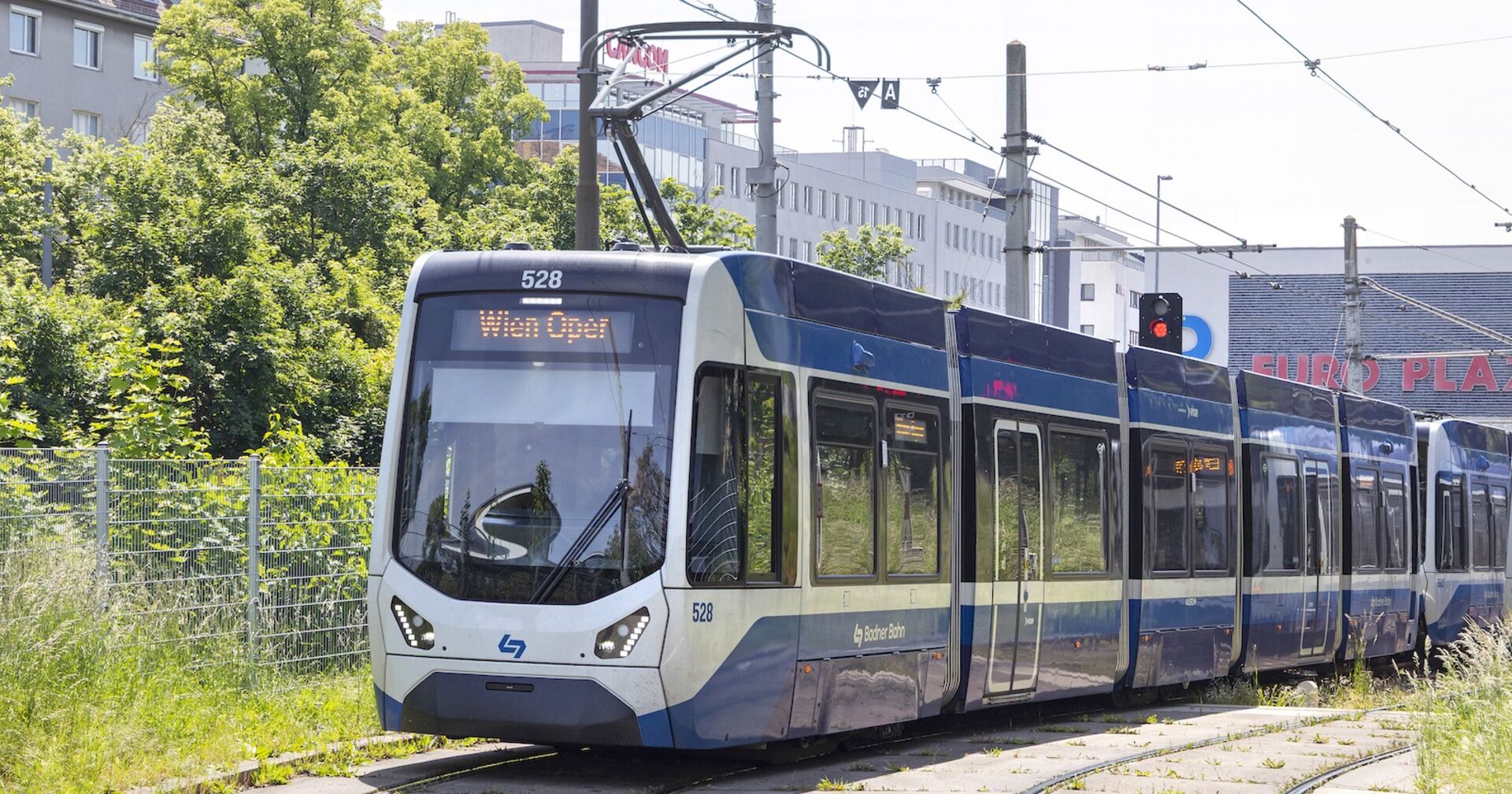 Moderne Garnitur der Badner Bahn mit Zielanzeige „Wien Oper“, fährt durch eine begrünte Gleisanlage vor städtischem Hintergrund, aufgenommen bei Tageslicht.