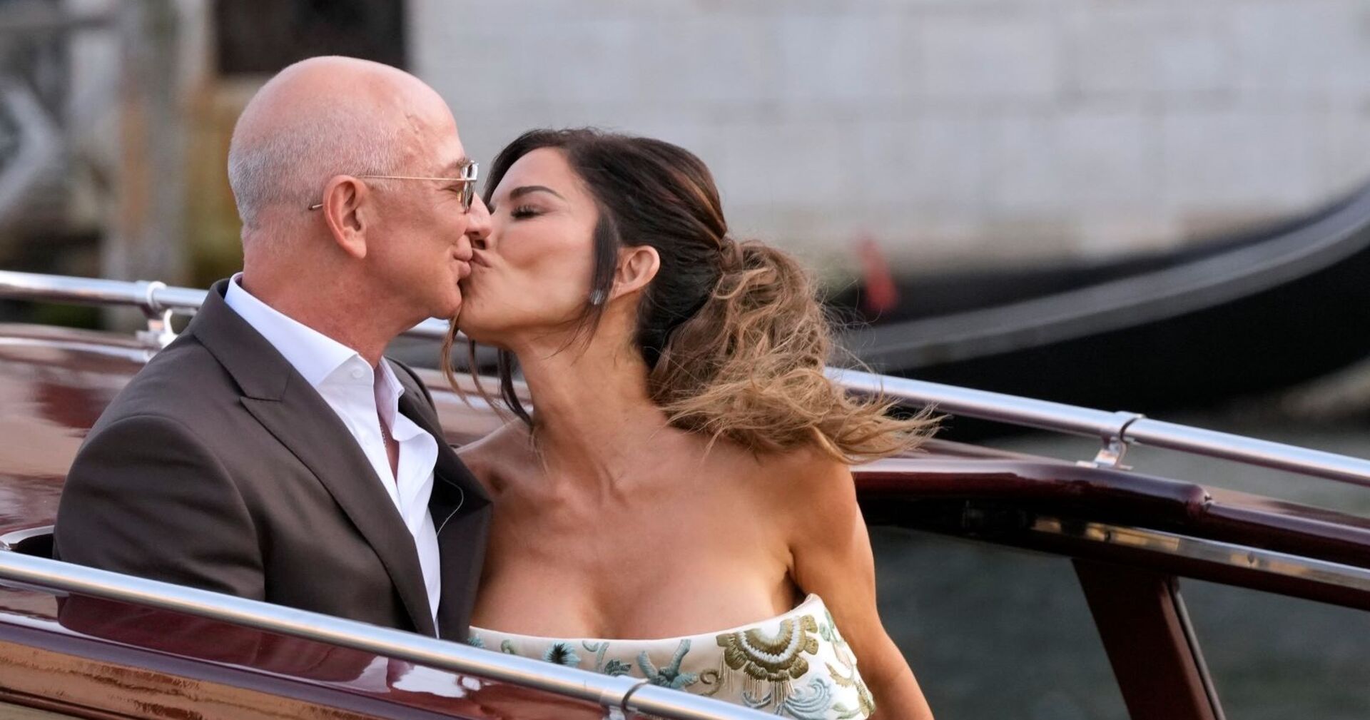 Jeff Bezos und Lauren Sánchez küssen sich innig auf einem Boot in Venedig; er trägt einen dunklen Anzug, sie ein schulterfreies Kleid mit Blumenmuster, im Hintergrund verschwimmt eine Gondel.