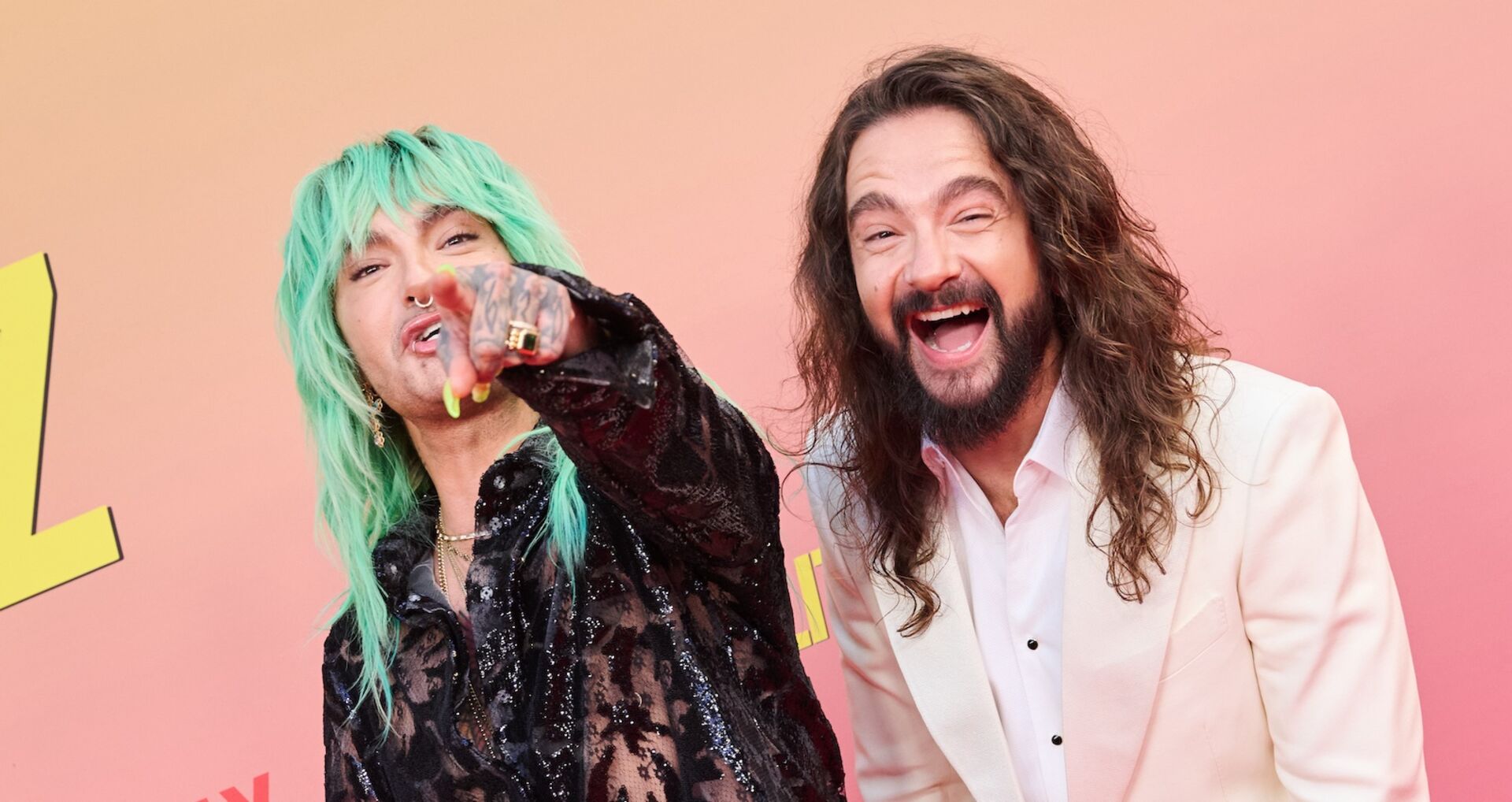 Bill Kaulitz mit neongrüner Frisur und durchsichtiger schwarzer Bluse zeigt lachend auf die Kamera, neben ihm steht Tom Kaulitz in weißem Anzug, beide posieren fröhlich vor rotem Hintergrund.