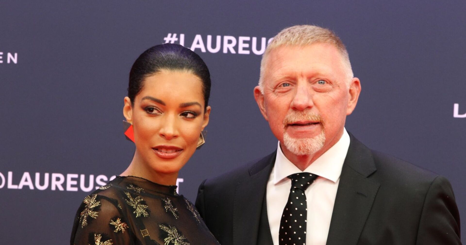 Boris Becker im schwarzen Anzug mit gepunkteter Krawatte und seine Frau Lilian de Carvalho Monteiro in einem schwarzen Kleid mit goldenen Details posieren gemeinsam auf dem roten Teppich