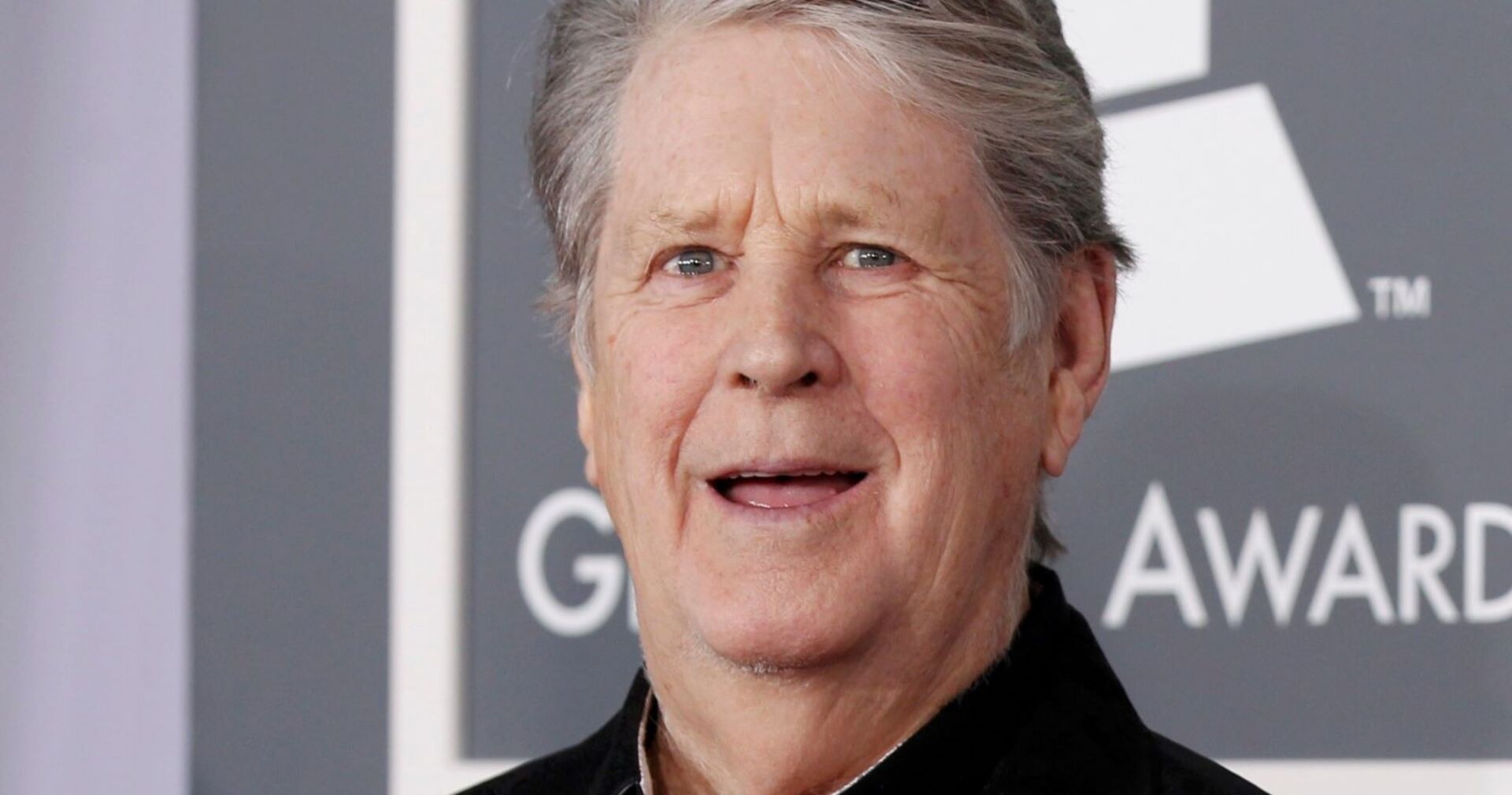 Porträt von Brian Wilson, lächelnd vor einem Grammy-Hintergrund, in fortgeschrittenem Alter.