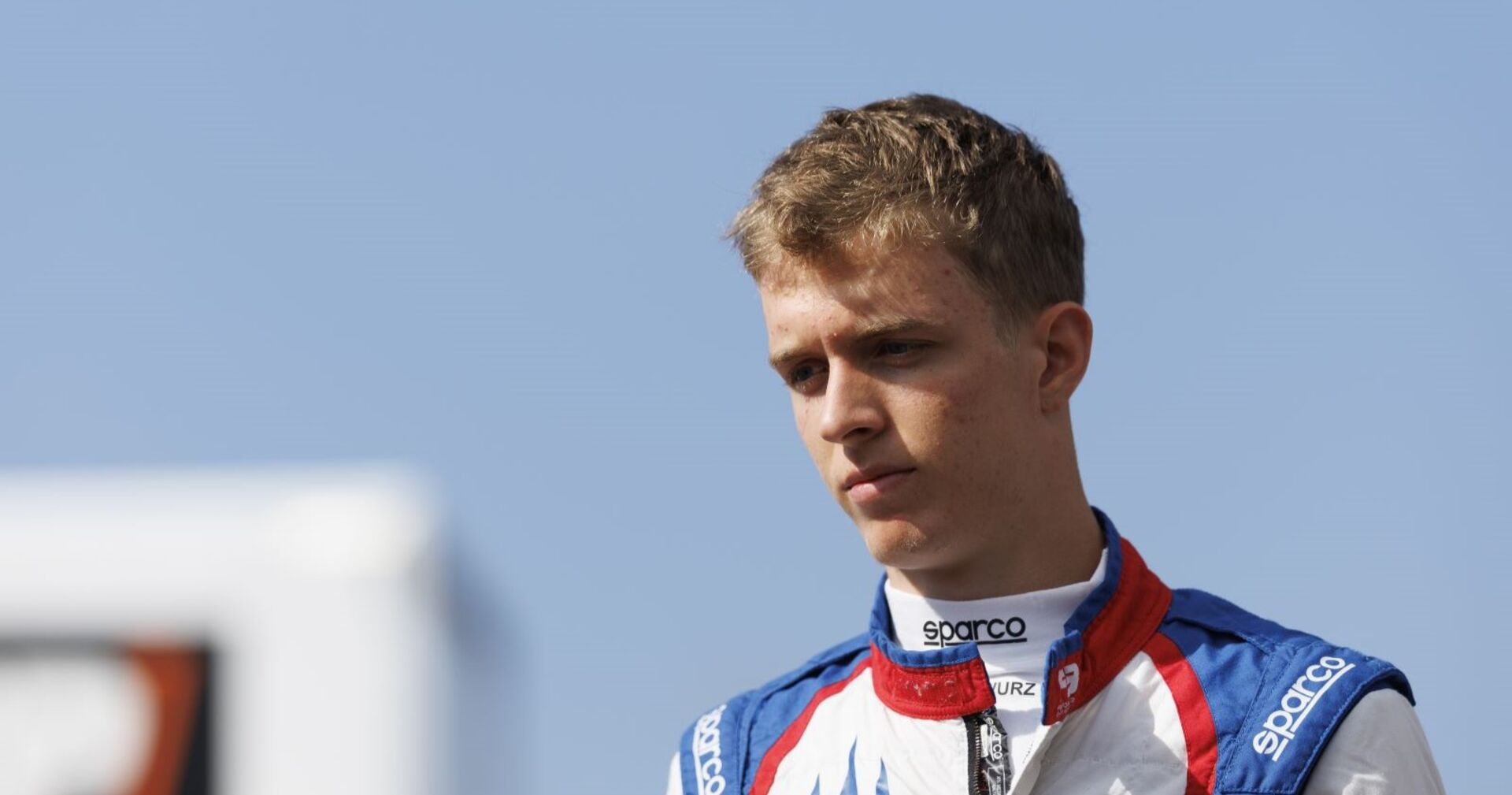 Charlie Wurz in Rennanzug von Sparco, blickt ernst nach unten, blauer Himmel im Hintergrund.