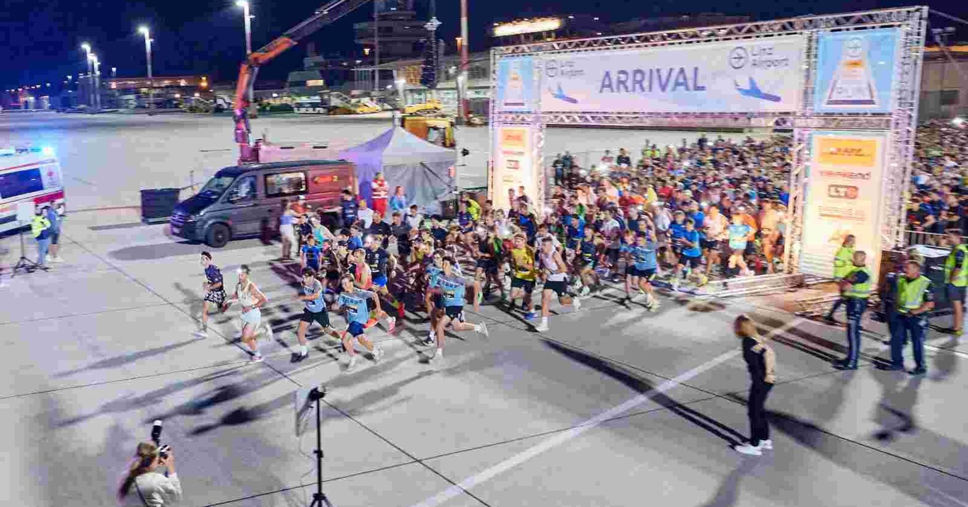 Eine große Gruppe von Läuferinnen und Läufern startet bei einem nächtlichen Lauf-Event auf dem Rollfeld eines Flughafens. Im Vordergrund sind die schnellsten Teilnehmenden bereits losgelaufen. Der Startbereich ist mit einem großen Torbogen mit der Aufschrift „ARRIVAL“ markiert. Rundherum stehen Zuschauende, Ordner in Warnwesten und Fotografen. Links im Bild befinden sich ein Krankenwagen und ein schwarzer Transporter, rechts sind Sponsorenbanner am Starttor zu sehen.