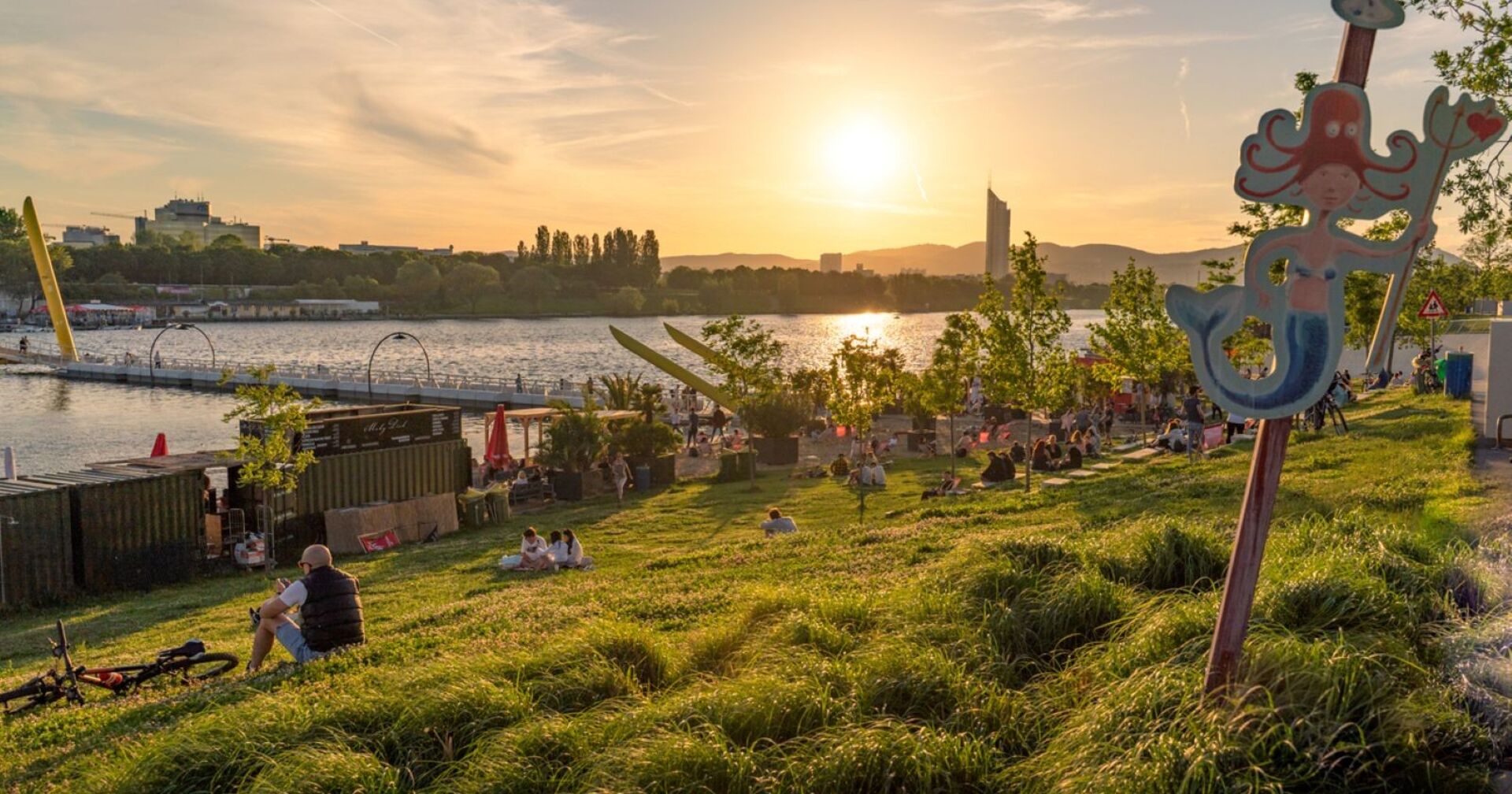 alte Donau im Sommer, Menschen sitzen auf Wiese am Wasser und beobachten den Sonnenuntergang
