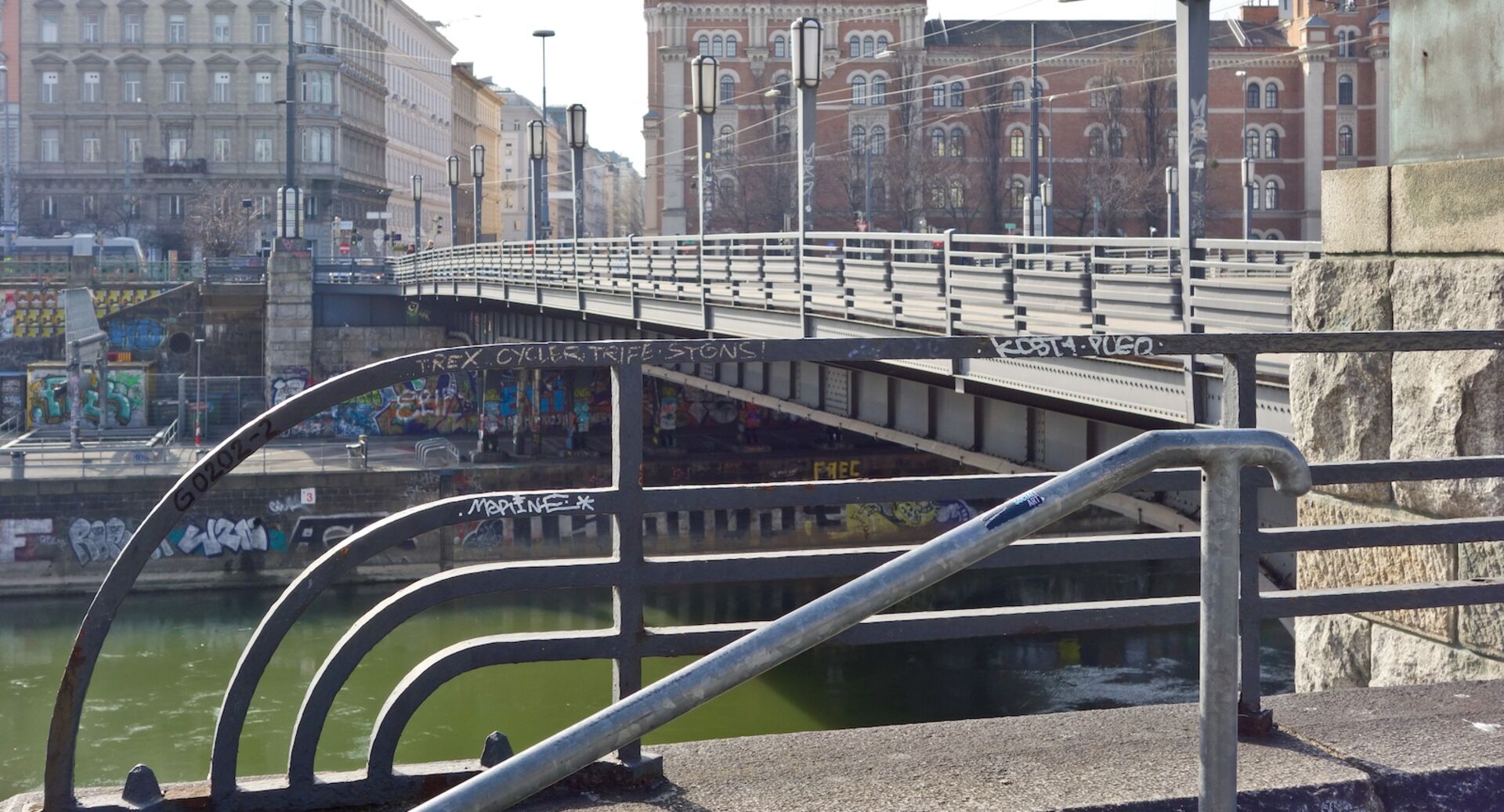 Blick auf die Augartenbrücke in Wien mit Blick auf den Donaukanal und Graffiti an den Ufermauern; im Vordergrund ein Geländer mit Schmierereien, im Hintergrund historische Gebäude.