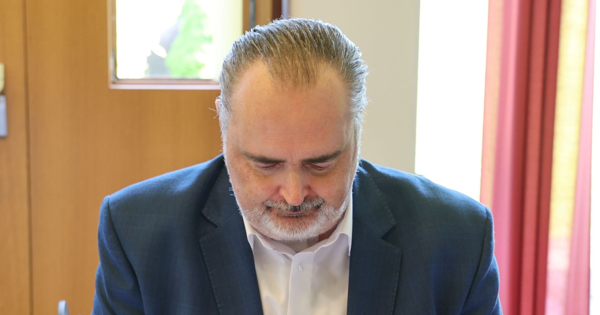 Hans Peter Doskozil blickt nach unten, trägt ein dunkles Sakko und ein weißes Hemd, im Hintergrund ein Fenster mit rotem Vorhang.