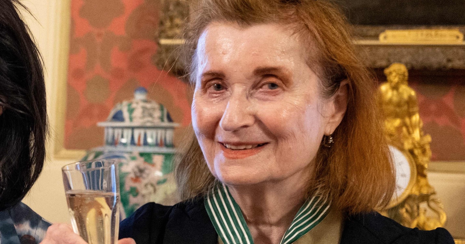 Elfriede Jelinek mit einem Glas in der Hand bei einer feierlichen Veranstaltung, freundlich lächelnd, im festlich dekorierten Innenraum