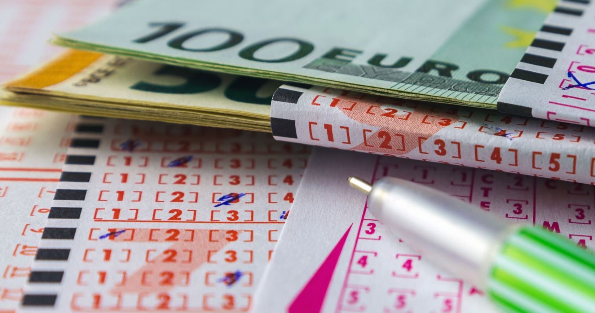 Nahaufnahme eines ausgefüllten Lottoscheins mit mehreren Euro-Geldscheinen und einem grünen Kugelschreiber.