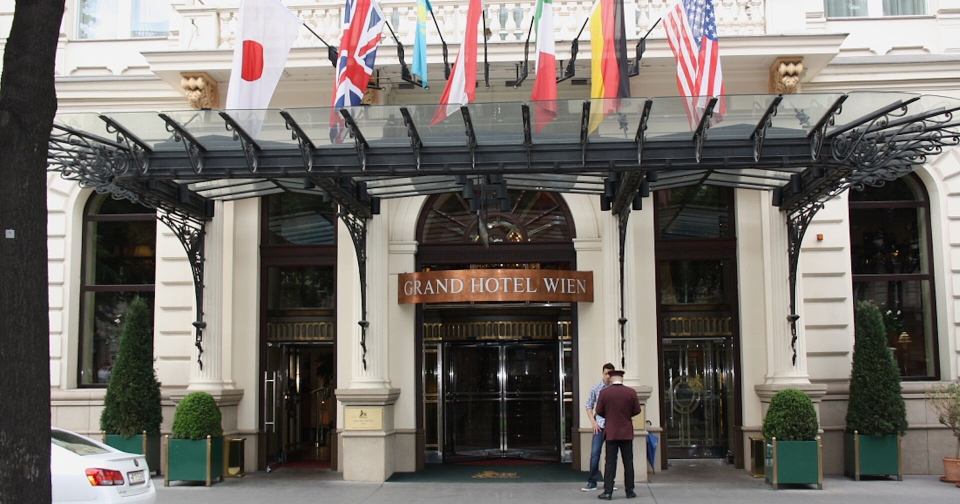 Eingang des Grand Hotel Wien mit Glasvordach, verzierten Eisenstützen und mehreren internationalen Flaggen, zwei Personen stehen vor dem Hoteleingang.