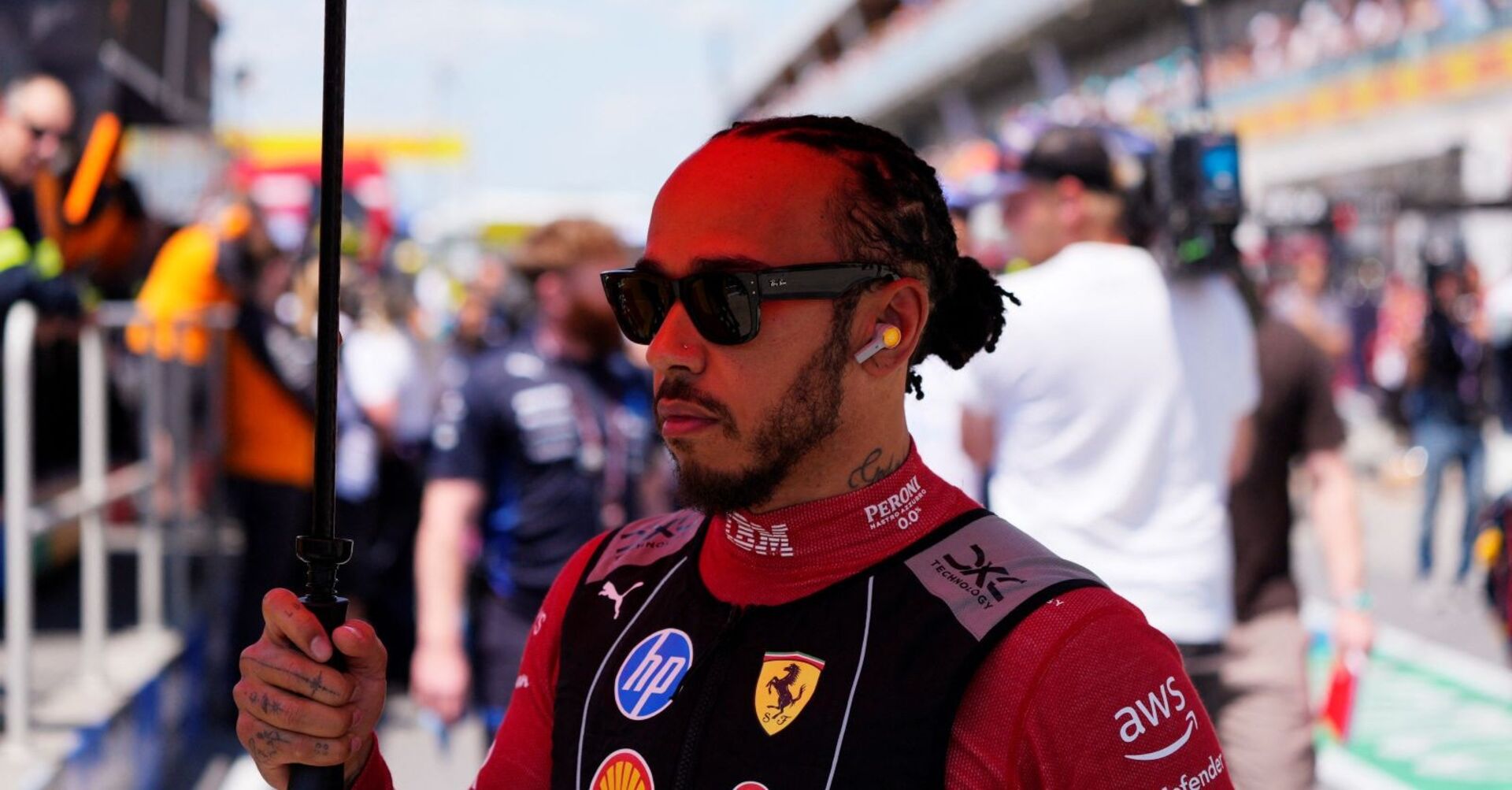 Lewis Hamilton im Ferrari-Rennoverall mit Sonnenbrille und Kopfhörern in der Startaufstellung des Kanada-GP 2025, hält einen Sonnenschirm, Hintergrund unscharf.
