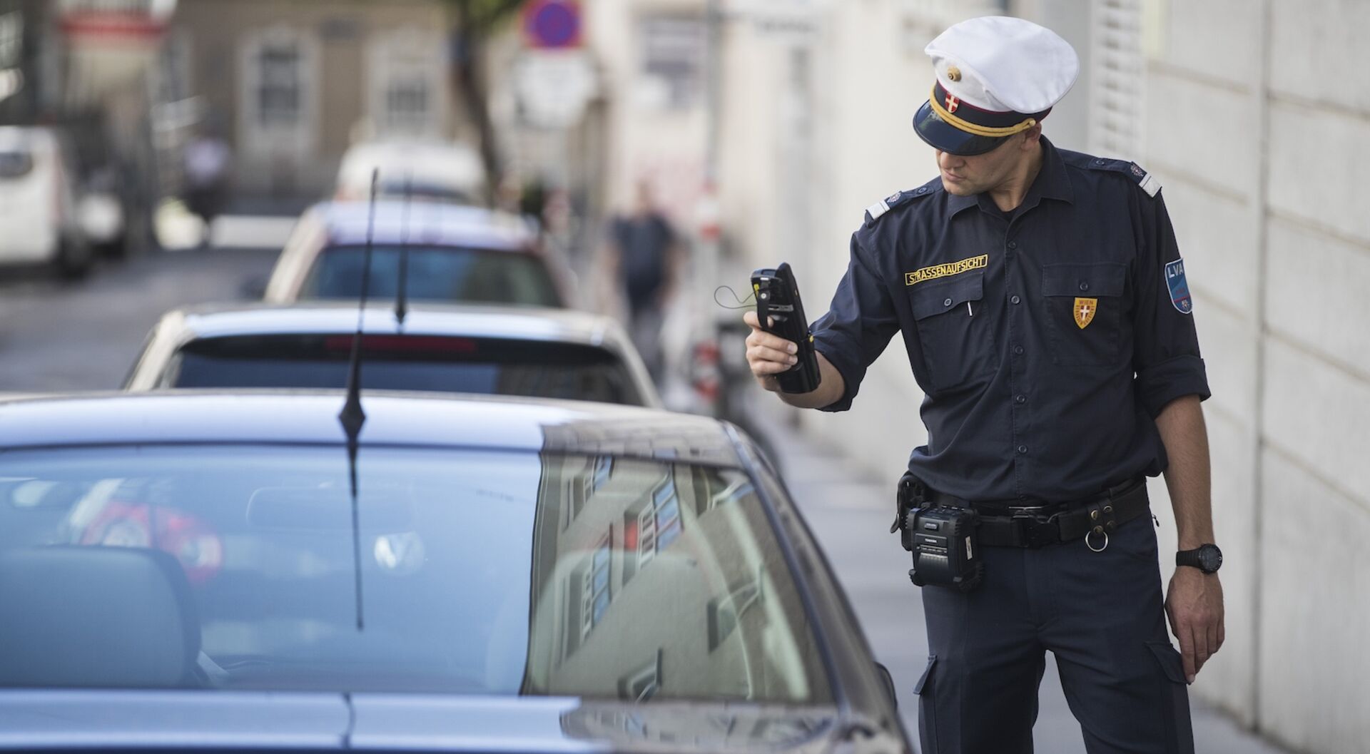 Straßenaufsichtsbeamter in Uniform scannt den Parkschein eines parkenden Auto in Wien mit einem mobilen Kontrollgerät.
