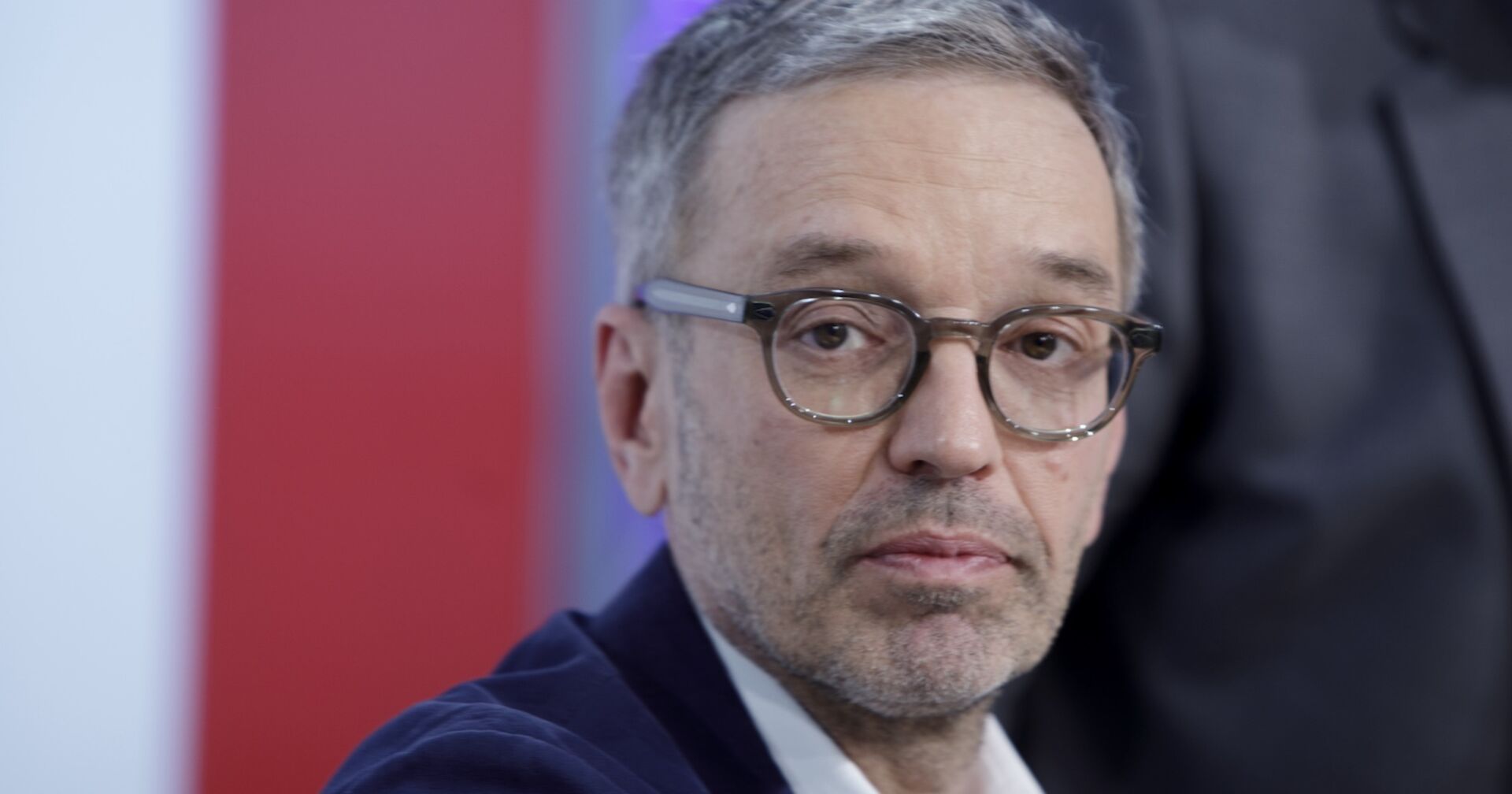 Herbert Kickl mit ernstem Gesichtsausdruck, trägt Brille und Sakko, vor rotem und weißem Hintergrund.