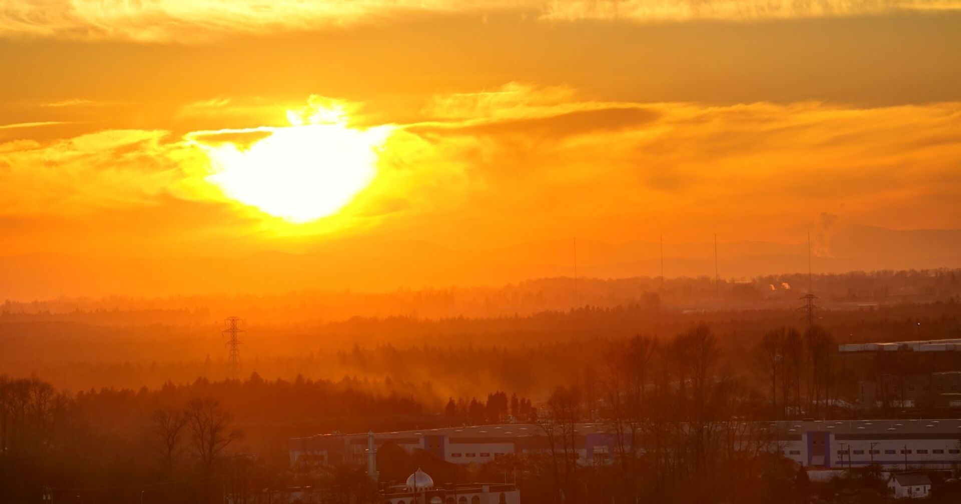 Sonnenuntergang mit glutrotem Himmel über einer bewaldeten Landschaft und Industriegebäuden, Hitze- und Dunstschleier sichtbar.