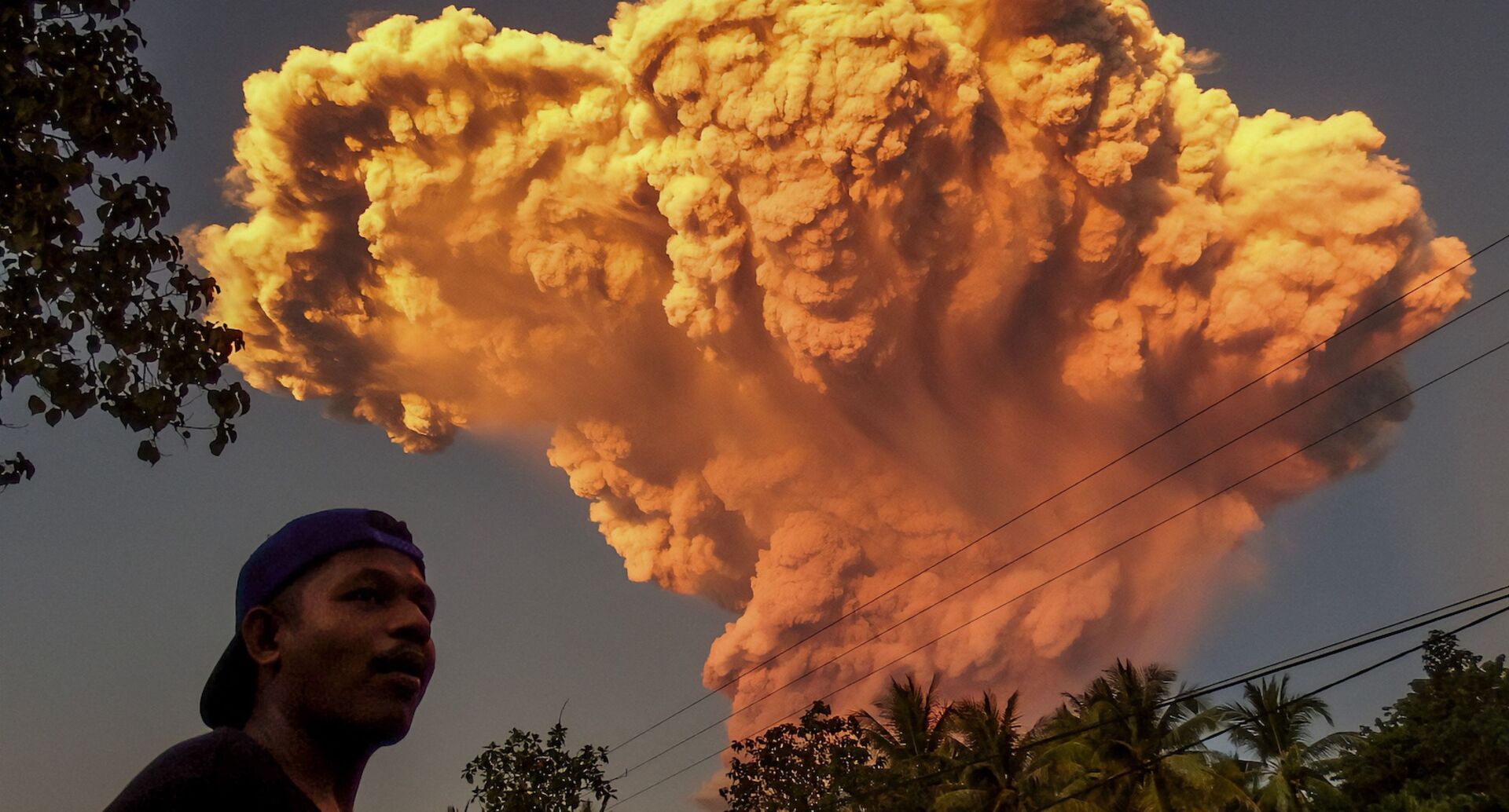 Ein Mann steht im Vordergrund, während im Hintergrund eine gewaltige orangefarbene Aschewolke vom ausbrechenden Vulkan Lewotobi Laki-Laki auf der indonesischen Insel Flores aufsteigt.