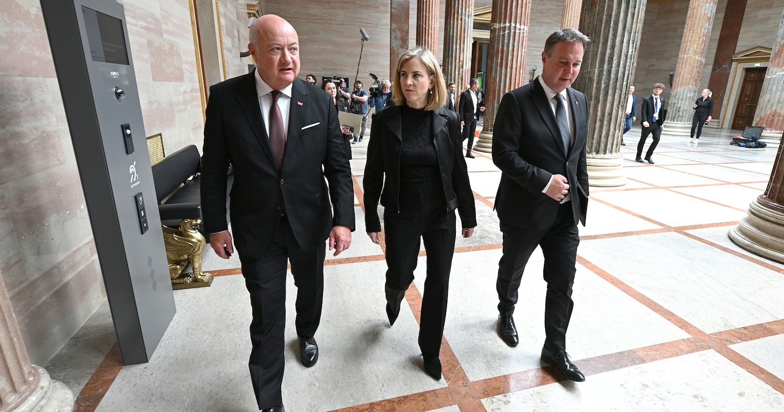 Bundeskanzler Christian Stocker, Außenministerin Beate Meinl-Reisinger und Vizekanzler Andreas Babler – gehen durch die Säulenhalle des Parlamentsgebäudes in Wien, begleitet von Medienvertretern.