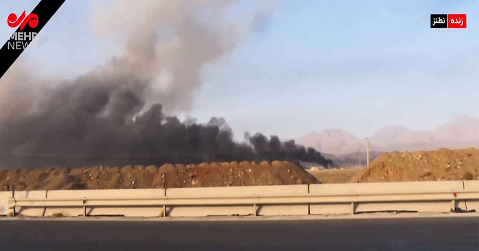 Standbild eines iranischen Fernsehsenders zeigt dunkle Rauchschwaden, die hinter einem Erdhügel und einer Straße aufsteigen. Im Hintergrund sind Berge zu sehen. Die Aufnahme stammt offenbar aus der Nähe der Anlage in Natans nach israelischen Luftschlägen.