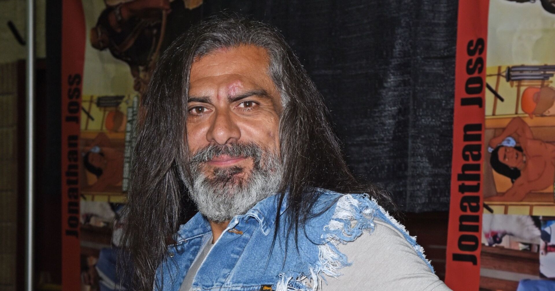Porträt von Jonathan Joss mit langen Haaren und Bart, im Jeanshemd vor einem Banner mit seinem Namen und einer Illustration seiner Figur John Redcorn aus „King of the Hill“.