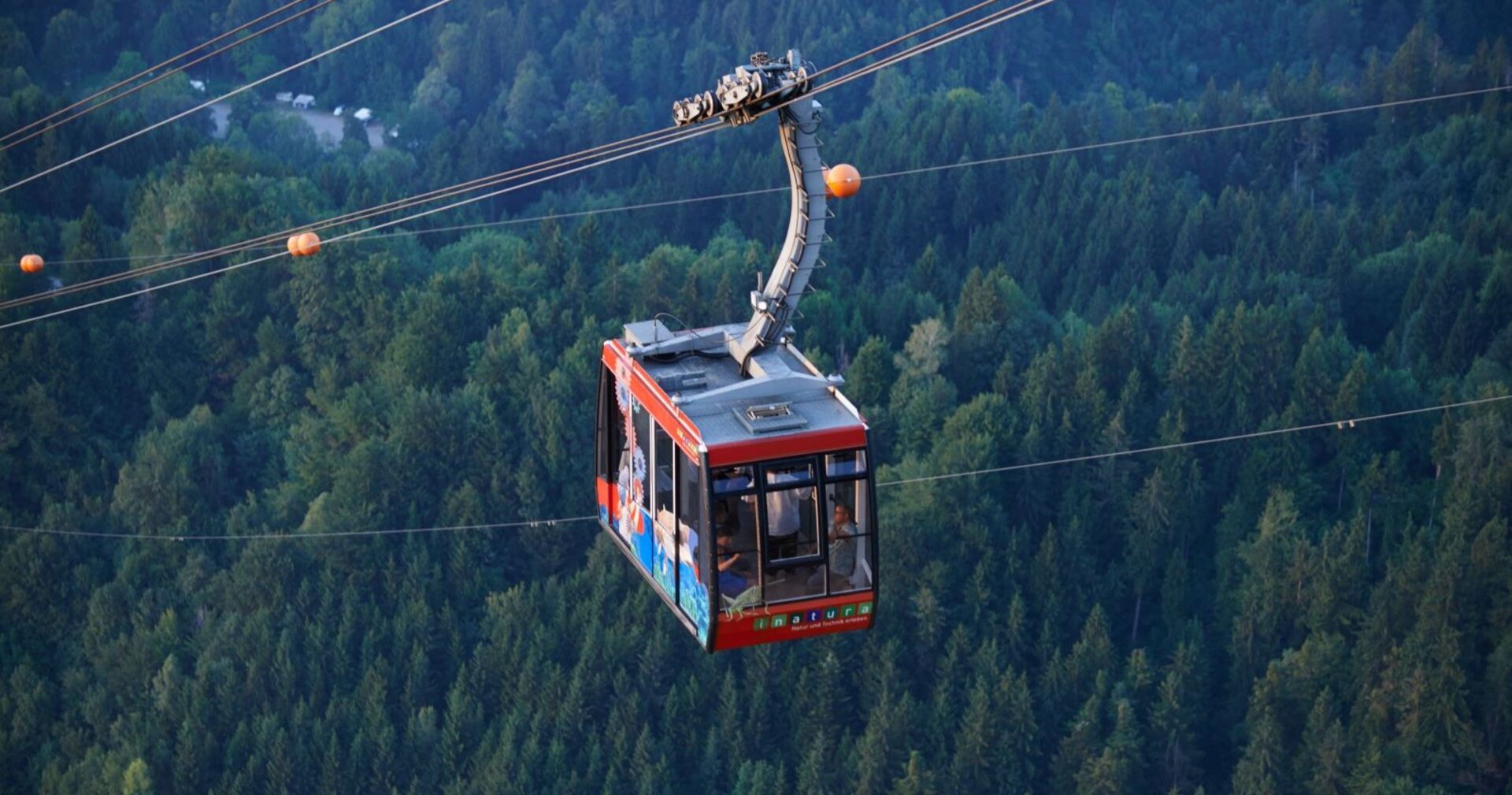 Eine rote Gondel der Karrenseilbahn hängt in großer Höhe über einem bewaldeten Tal bei Dornbirn. In der Kabine sind mehrere Personen zu sehen.
