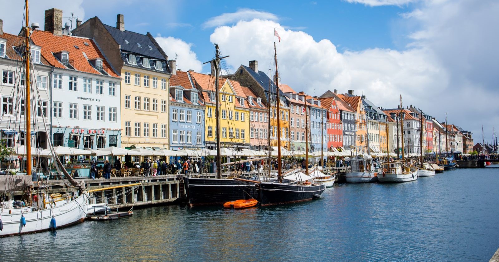 Bunte Giebelhäuser und historische Segelboote am Hafenkanal Nyhavn in Kopenhagen, bei Sonnenschein mit lebhaftem Treiben entlang der Promenade.