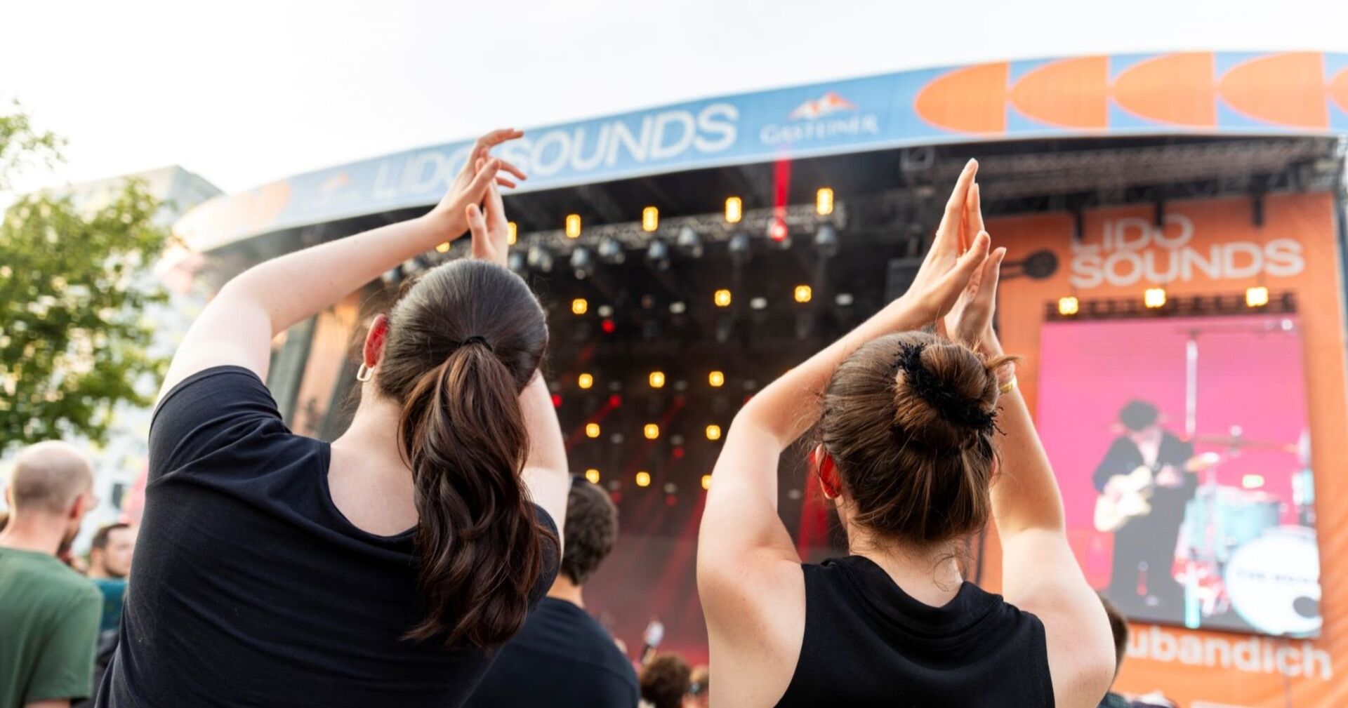 Zwei junge Frauen klatschen und feiern in einer Menschenmenge vor der Lido-Sounds-Bühne in Linz. Im Hintergrund sind bunte Lichter, ein LED-Screen mit Live-Musik und das Logo des Festivals zu sehen.