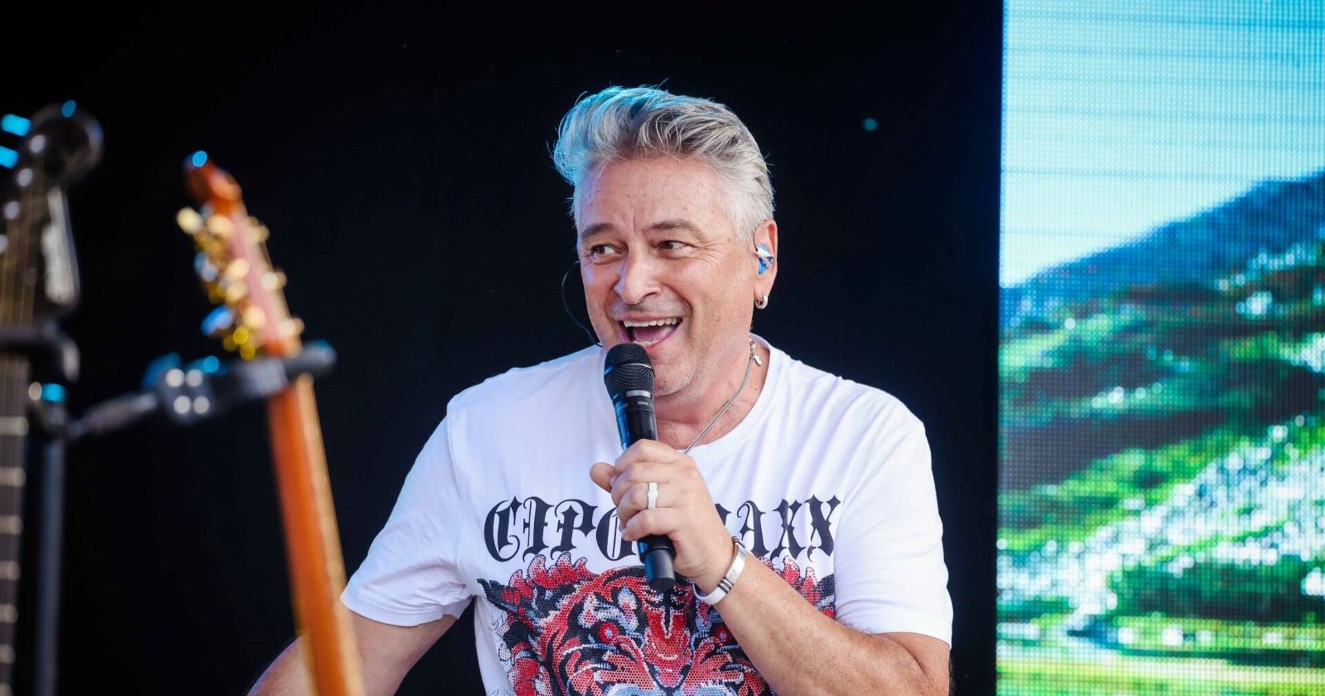 Didi Ganshofer, Sänger der Paldauer, mit Mikrofon auf der Bühne, lächelnd in einem weißen T-Shirt mit Tigerprint – Aufnahme vor seinem Schlaganfall.