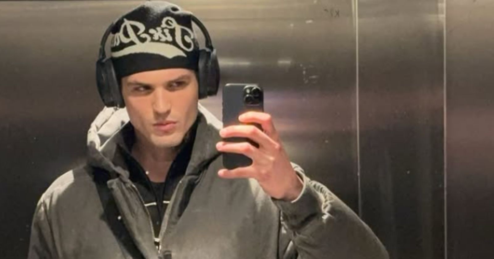 Pierre trägt grau-schwarz-weiße Kleidung, eine Jacke, eine Mütze und Kopfhörer und macht ein Spiegel-Selfie