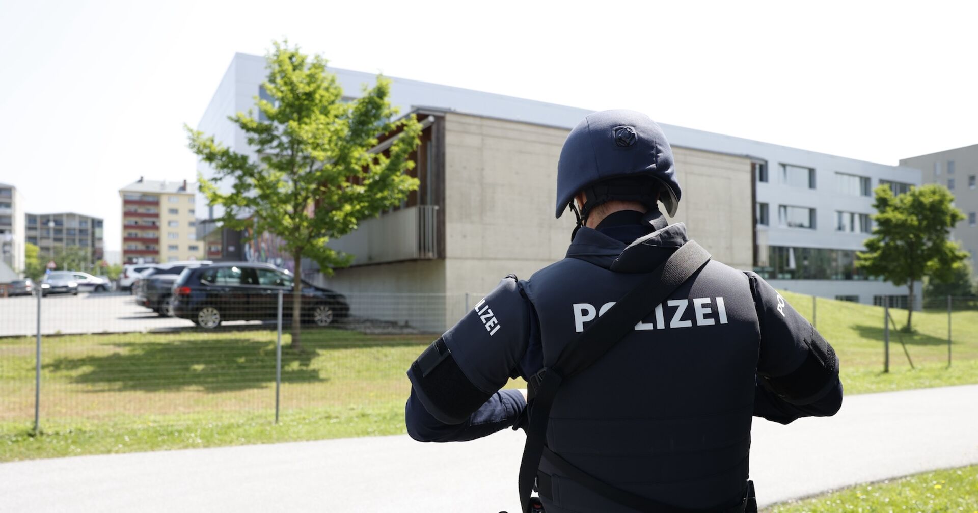 Ein Polizist in Schutzkleidung und mit Helm steht vor einem modernen Schulgebäude. Im Hintergrund sind parkende Autos und weitere Gebäude sichtbar. Die Szene spielt bei Tageslicht im Freien, das Gelände ist eingezäunt.