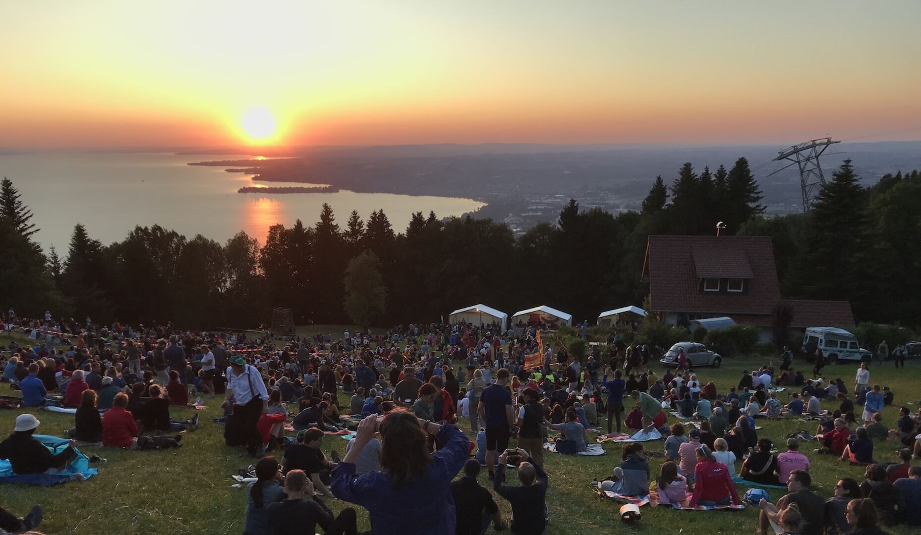 Menschen sitzen auf einer Wiese am Pfänder und blicken auf den Bodensee bei Sonnenuntergang während des Sonnwend-Fests mit Feuerstelle und Festzelten.