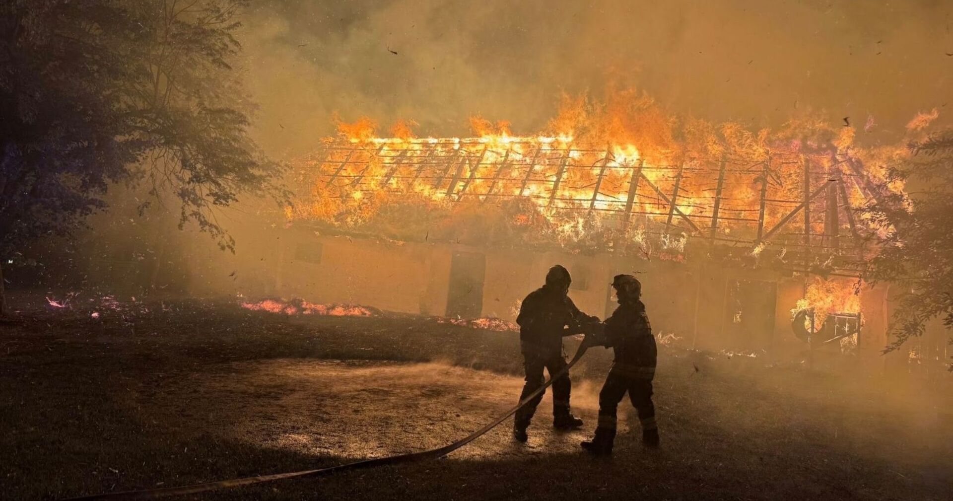 Zwei Feuerwehrleute bekämpfen in der Nacht einen Großbrand am Heimathaus Stinatz; das Strohdach steht lichterloh in Flammen.