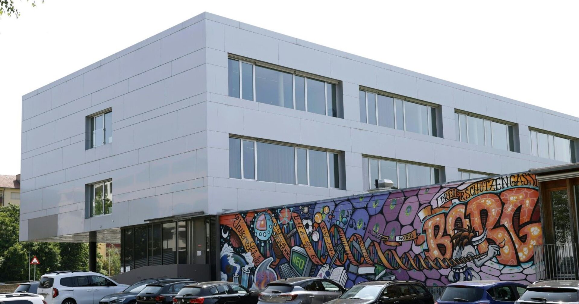 Modernes Schulgebäude mit weißer Fassade und großem Graffiti mit der Aufschrift „BORG Dreierschützengasse“ an der Frontwand. Mehrere Autos sind davor geparkt.
