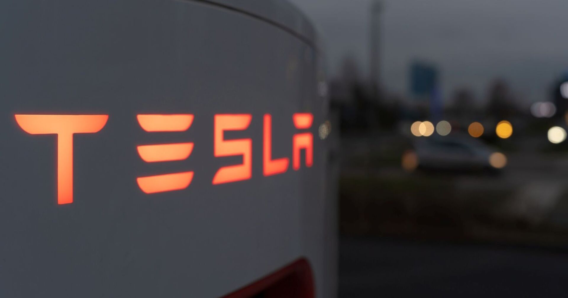Leuchtender Tesla-Schriftzug an einer Ladesäule bei Dämmerung, im Hintergrund unscharfe Straßenlichter und Autos.. Nach dem öffentlichen Streit zwischen Elon Musk und Donald Trump stürzt die Tesla-Aktie ab.